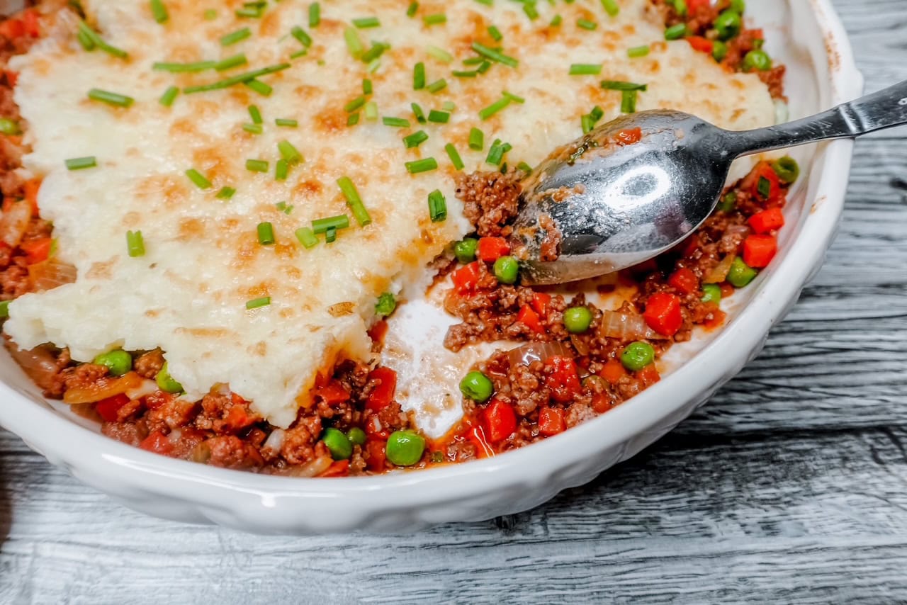 A close-up of a shepherd&rsquo;s pie in a white dish, showing a layer of mashed potatoes on top, garnished with chopped chives, and a meat and vegetable filling with peas and carrots visible where a serving spoon has scooped some out.
