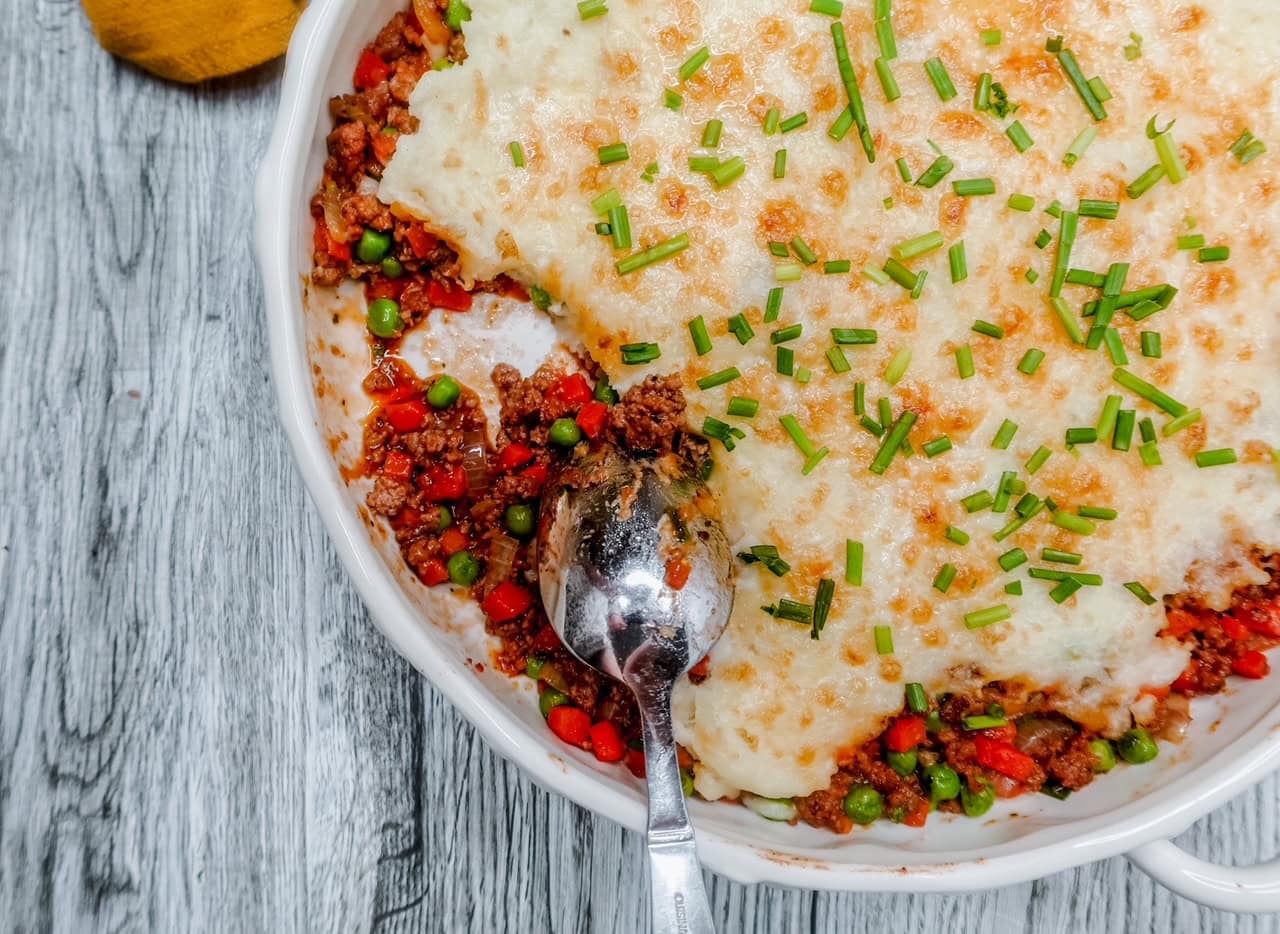 A close-up of a casserole dish of shepherd&rsquo;s pie with a serving missing, showing layers of ground meat, peas, and carrots topped with mashed potatoes and chopped chives, with a serving spoon resting in the dish.