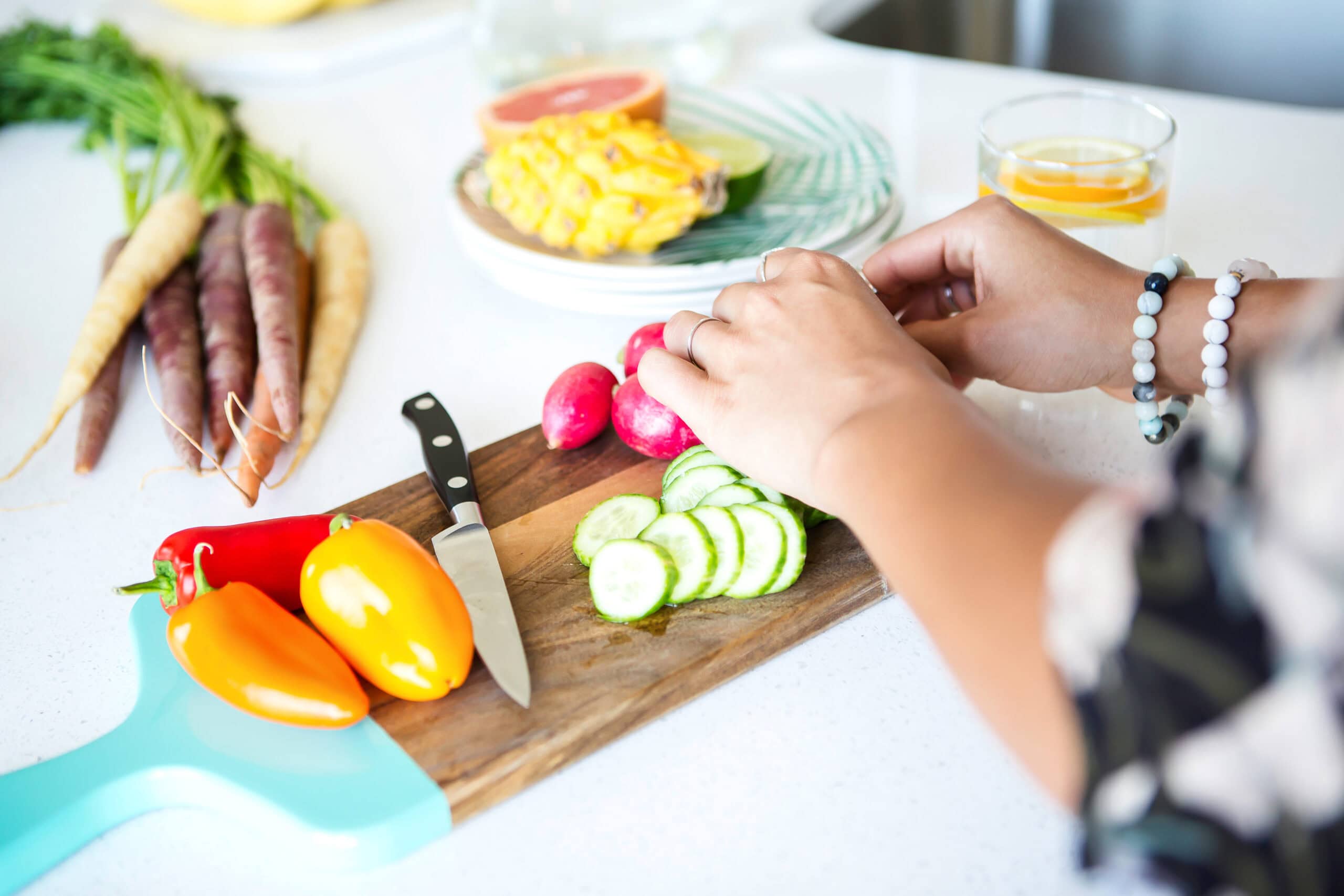 A person arranges cucumbers on a wooden cutting board surrounded by radishes, yellow and orange peppers, a knife, multicolored carrots, and a glass of water with orange slices on a white kitchen counter. There is a dish with fresh fruit blurred in the background.