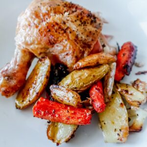 A serving of roasted chicken with crispy skin, accompanied by roasted potato wedges, carrots, and onions on a white plate. The vegetables appear golden and slightly caramelized.