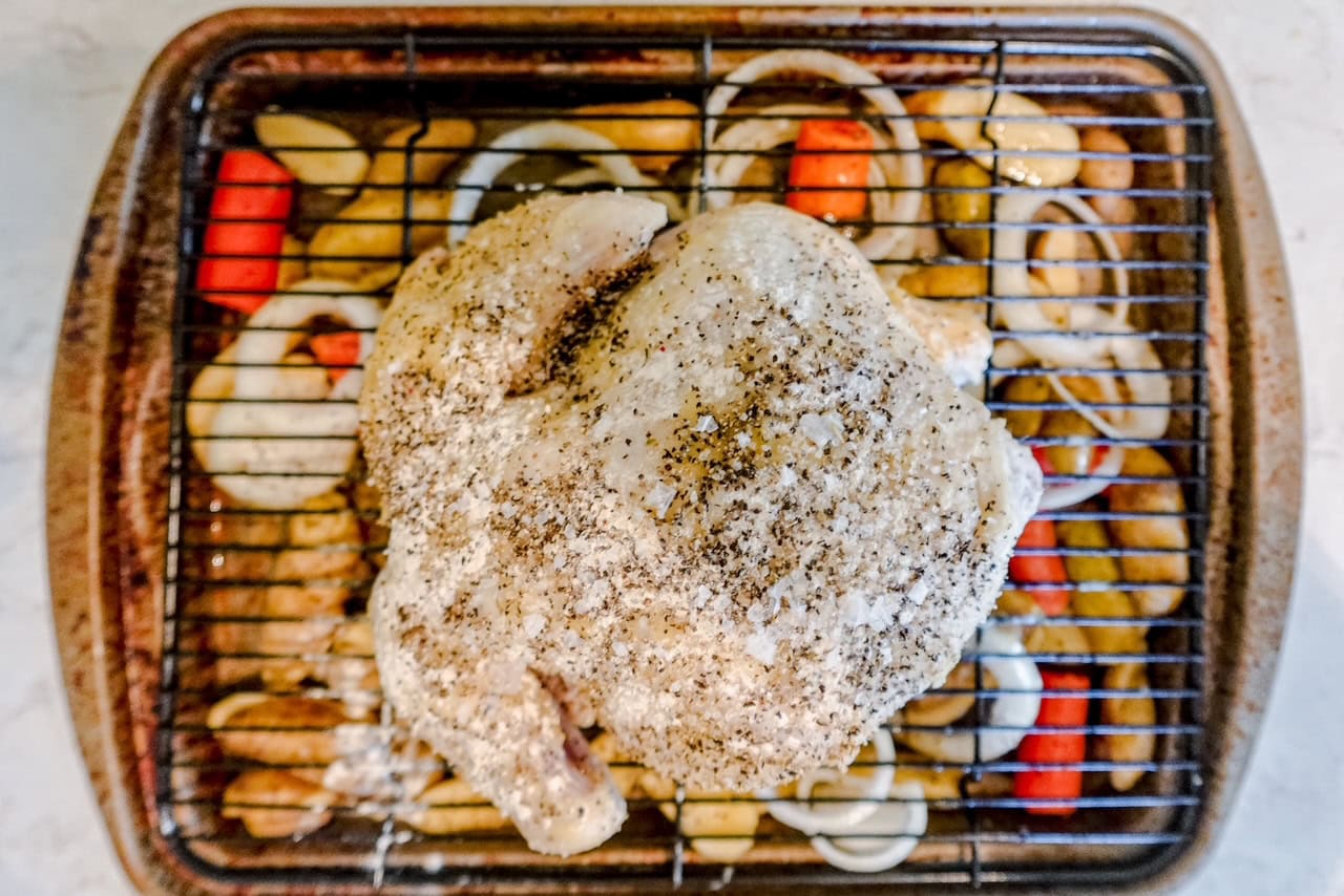 A seasoned whole raw chicken sits on a rack above sliced onions, red peppers, and potatoes in a roasting pan, ready to be cooked.