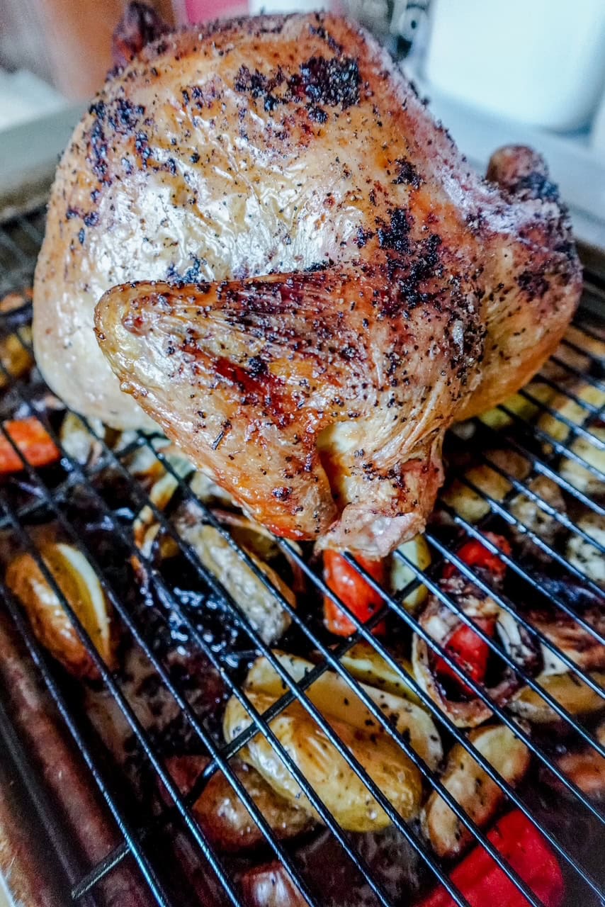 A whole roasted chicken sits on a wire rack above a tray of assorted roasted vegetables, including potatoes and carrots. The chicken is golden brown with crispy skin and seasoned with herbs.