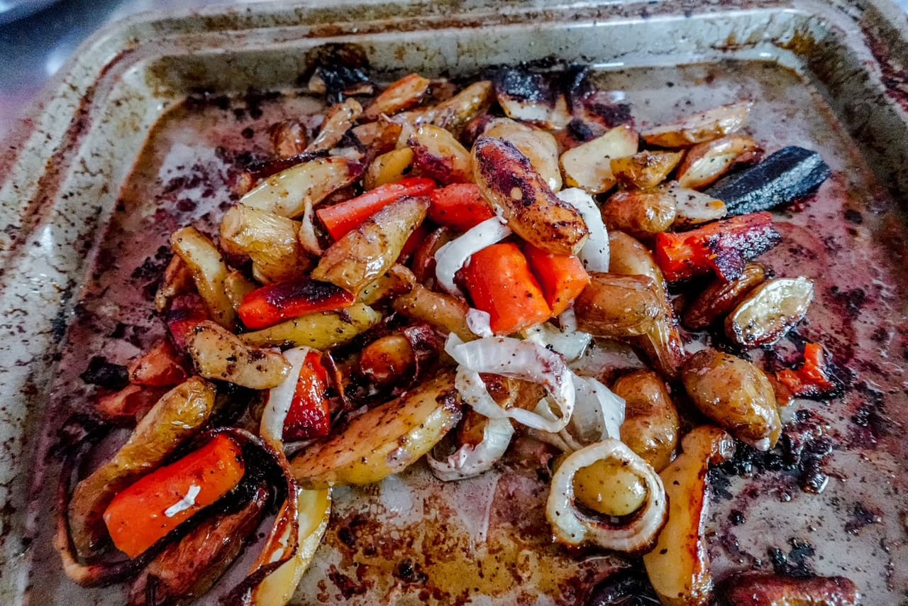 Roasted vegetables, including carrots, potatoes, and onions, are spread on a just-used baking tray. The vegetables are browned and slightly charred, indicating they have been roasted.