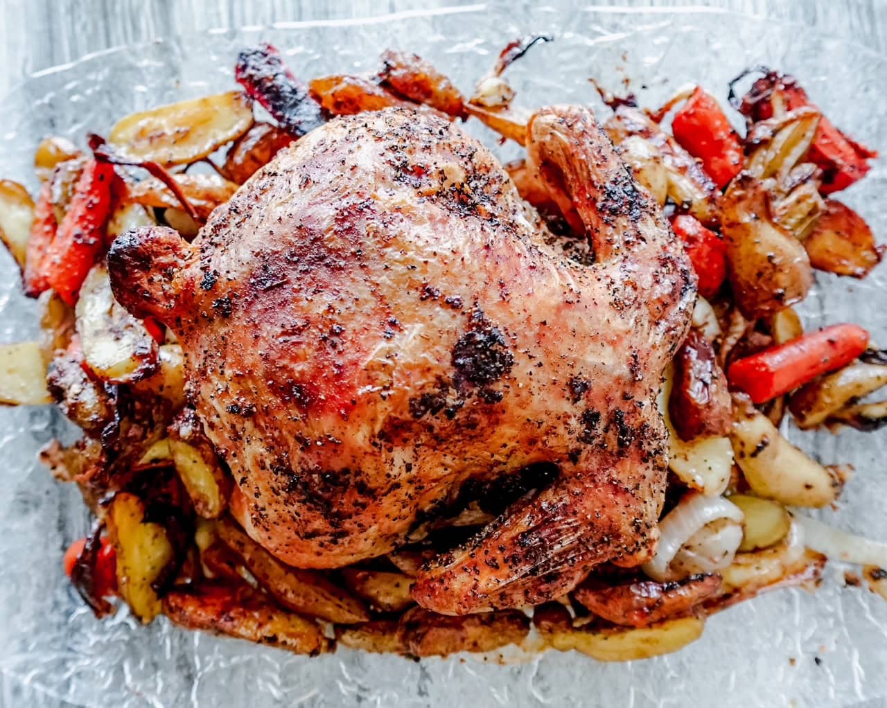 Overhead shot of a whole roasted chicken with crispy, seasoned skin sits on a bed of roasted vegetables, including potatoes, carrots, and onions, on a light gray background.