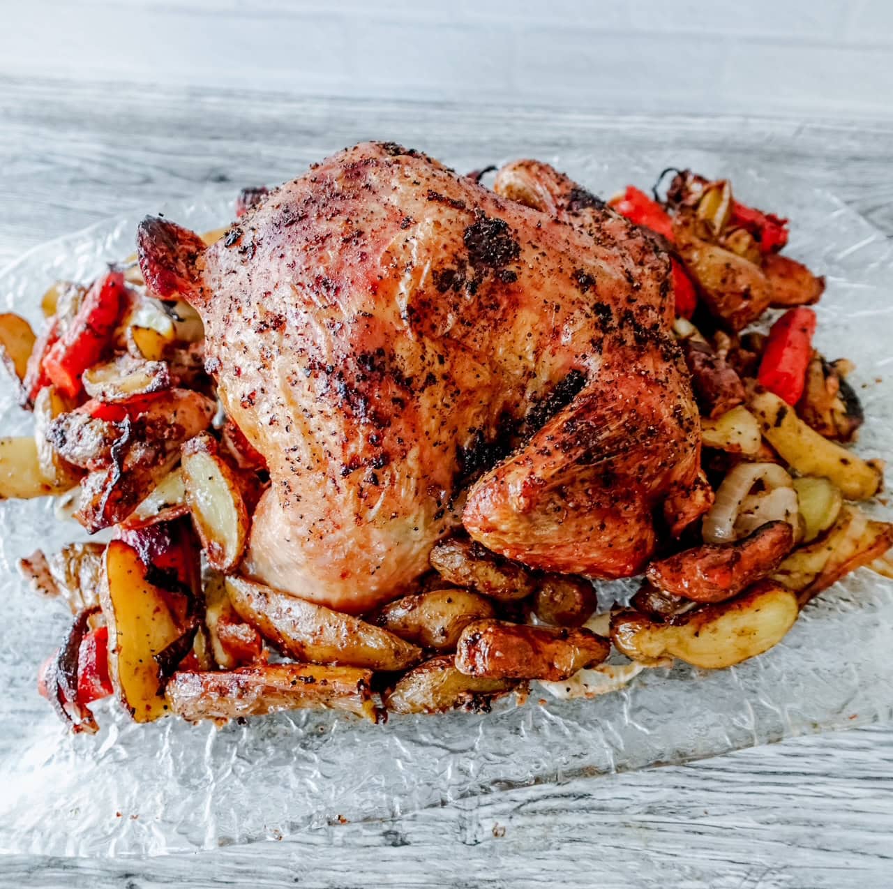 A whole roasted chicken with crispy skin is served on a bed of roasted potatoes, red bell peppers, and onions on a sheet of aluminum foil on a light gray wooden tabletop.