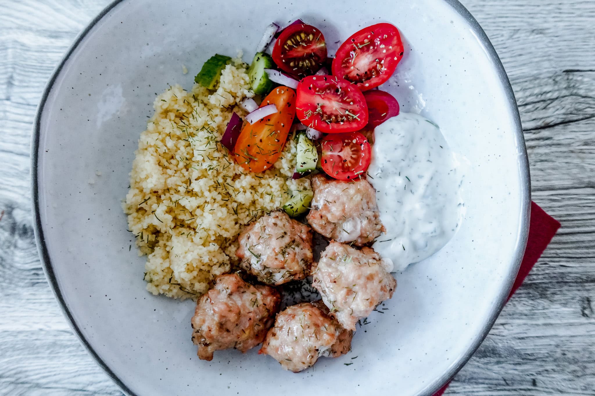 A bowl with baked chicken meatballs, couscous, sliced cherry tomatoes and cucumbers, red onion, and a serving of creamy tzatziki sauce, all arranged neatly on a white plate.