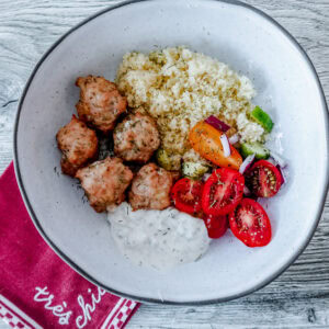 A bowl with Greek-style chicken meatballs, cooked couscous, grape tomatoes, cucumber, red onion, and a serving of creamy tzatziki sauce, placed on a red napkin on a wooden surface.