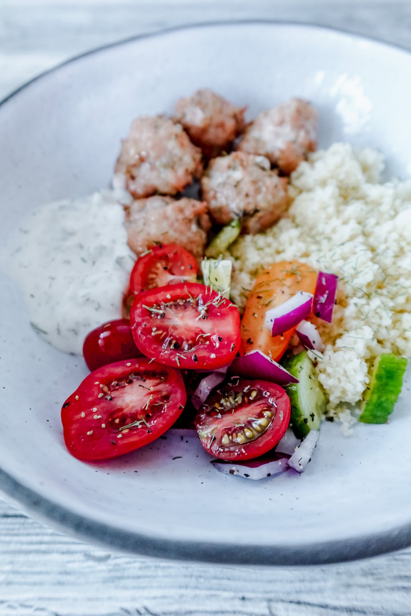 A bowl with Greek-style chicken meatballs, a serving of couscous, cherry tomato and cucumber salad with red onion, and a dollop of creamy tzatziki sauce, garnished with herbs.