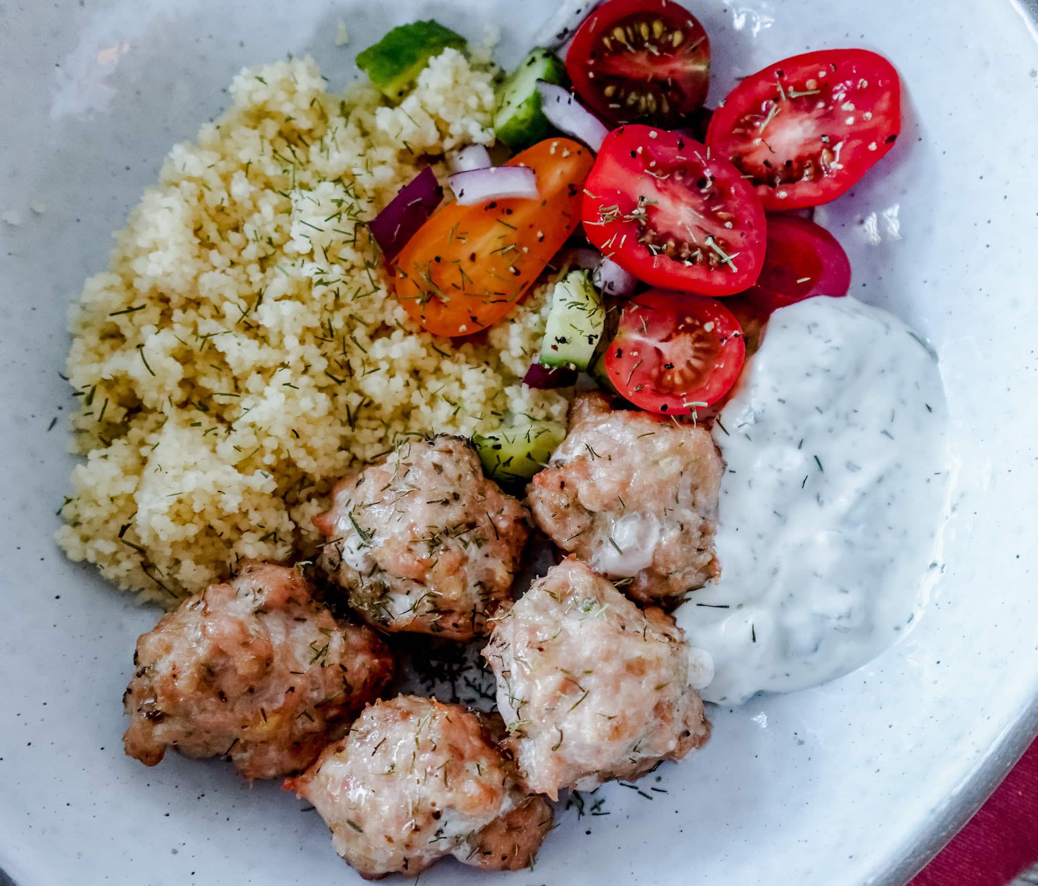 A plate with four meatballs, a serving of couscous, a colorful salad with tomatoes, cucumber, and red onion, and a dollop of creamy tzatziki sauce.