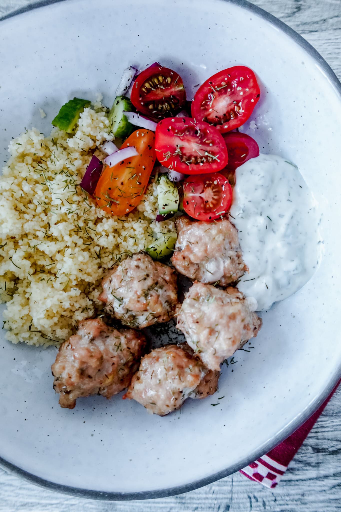 A white bowl with baked chicken meatballs, a scoop of couscous, sliced cherry tomatoes, cucumbers, red onions, carrots, and a serving of creamy white tzatziki sauce.