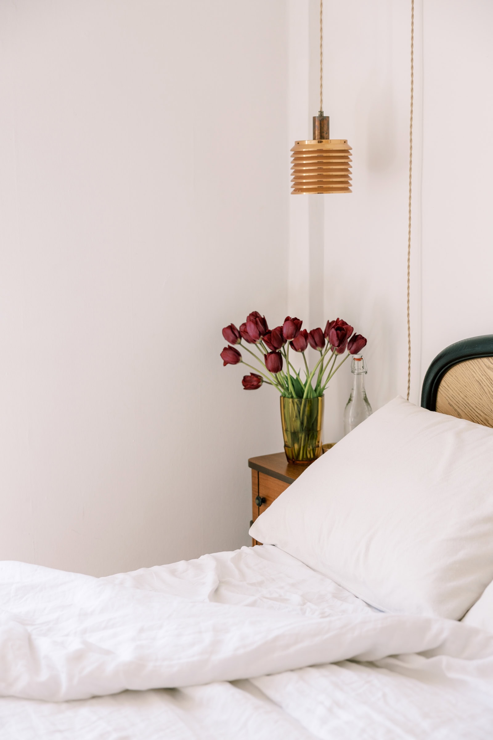 A cozy bedroom with white bedding, a pillow, a bedside table holding a vase of dark red tulips, and a small hanging wooden lamp against a white wall.
