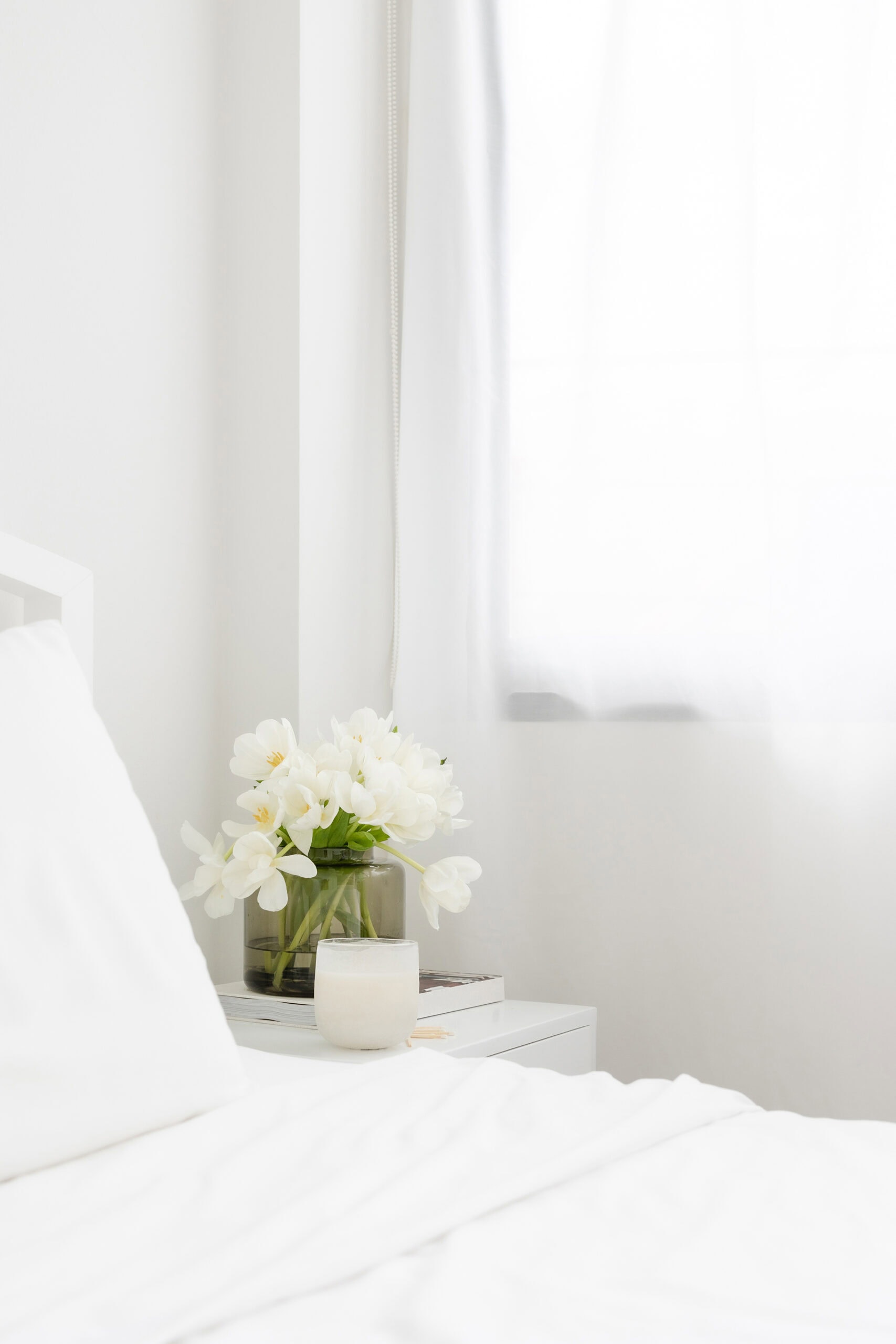 A bright, minimalist bedroom with white bedding, a bedside table holding a vase of white flowers, a candle, and a book, next to a window with sheer white curtains letting in soft natural light.
