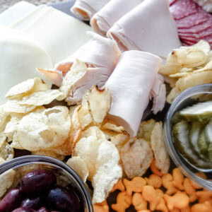 A close-up of a charcuterie board featuring rolled deli meats, sliced cheese, salami, potato chips, goldfish crackers, pickles in a small dish, and a bowl of black olives.