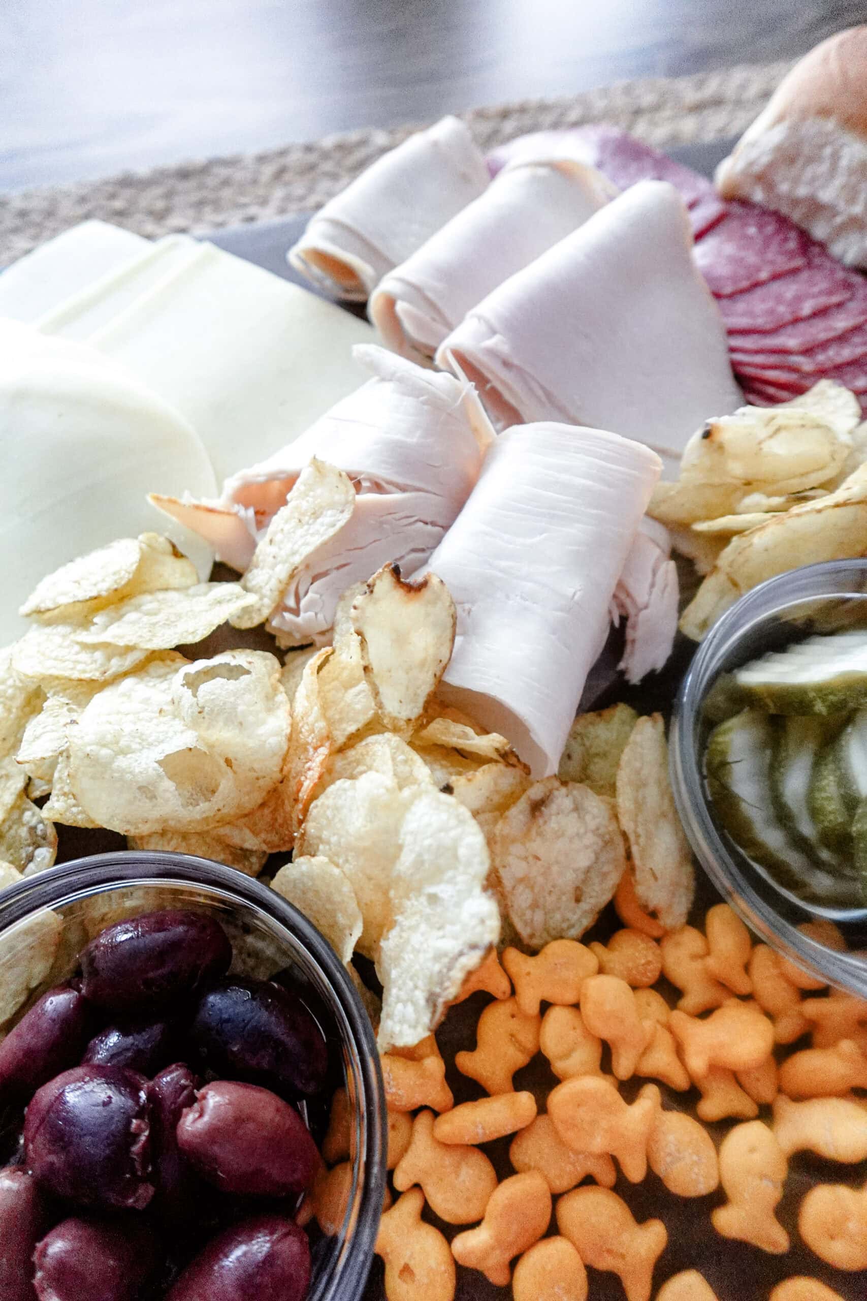 A close-up of a charcuterie board featuring rolled deli meats, sliced cheese, salami, potato chips, goldfish crackers, pickles in a small dish, and a bowl of black olives.