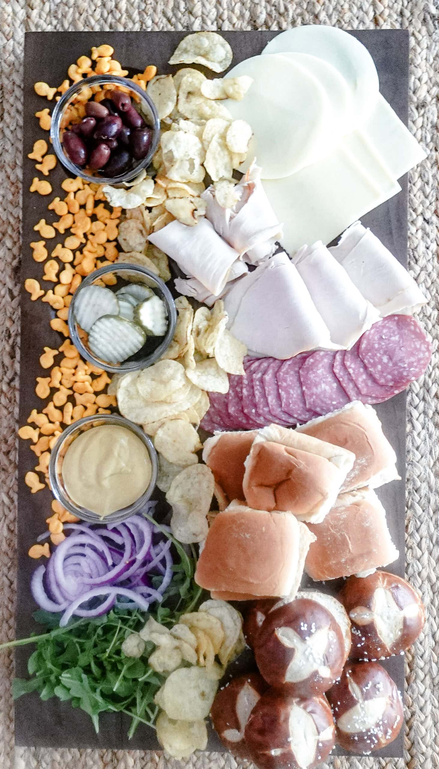 An overhead shot of a charcuterie board with sliders, sliced salami, turkey, provolone cheese, pickles, red onion, arugula, potato chips, mustard, Goldfish crackers, olives, and pretzel rolls on a woven mat.
