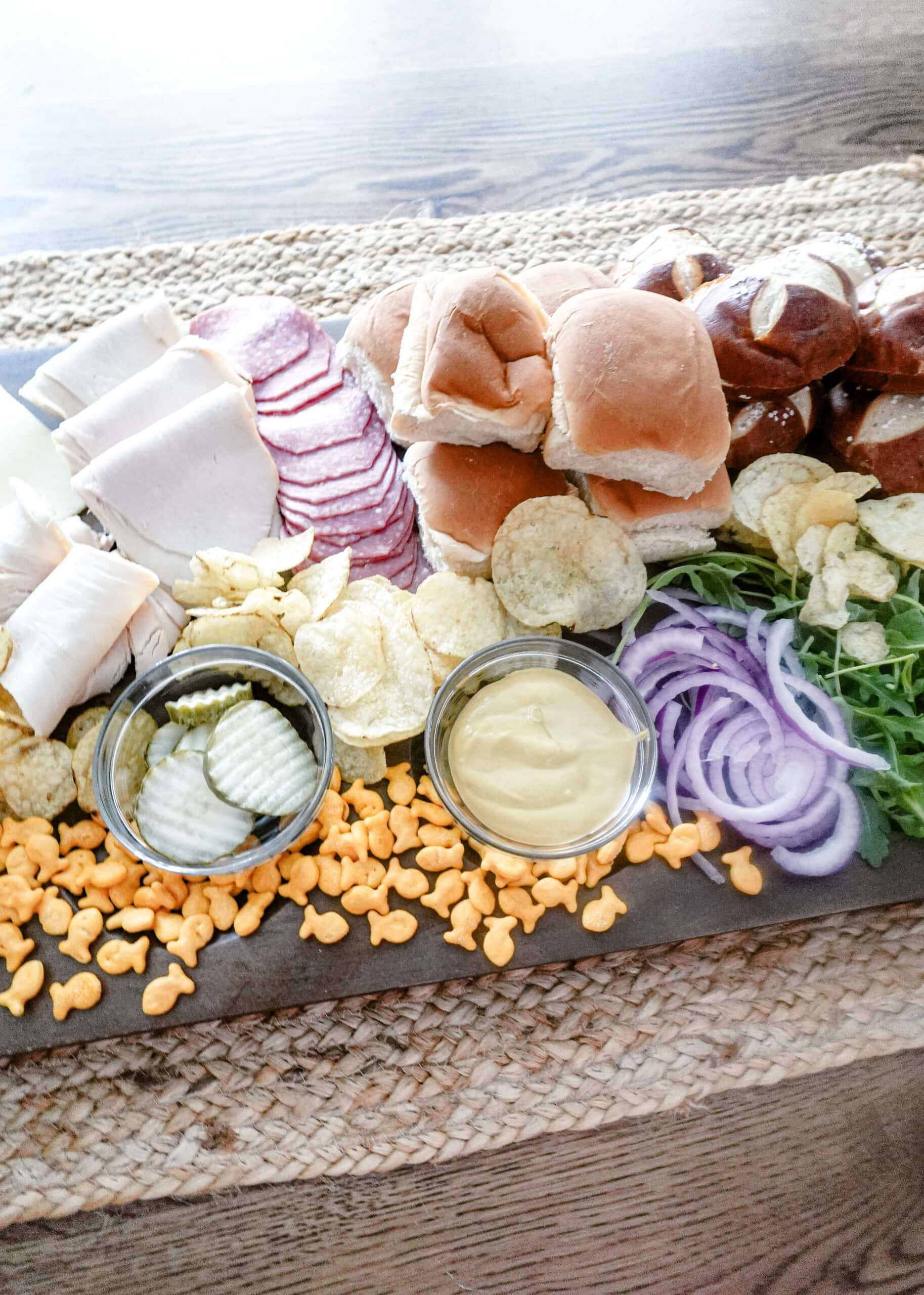 An overhead shot of a charcuterie board with sliced deli meats, pretzel and slider buns, potato chips, Goldfish crackers, pickle slices, mustard, sliced red onions, and fresh greens on a woven placemat.