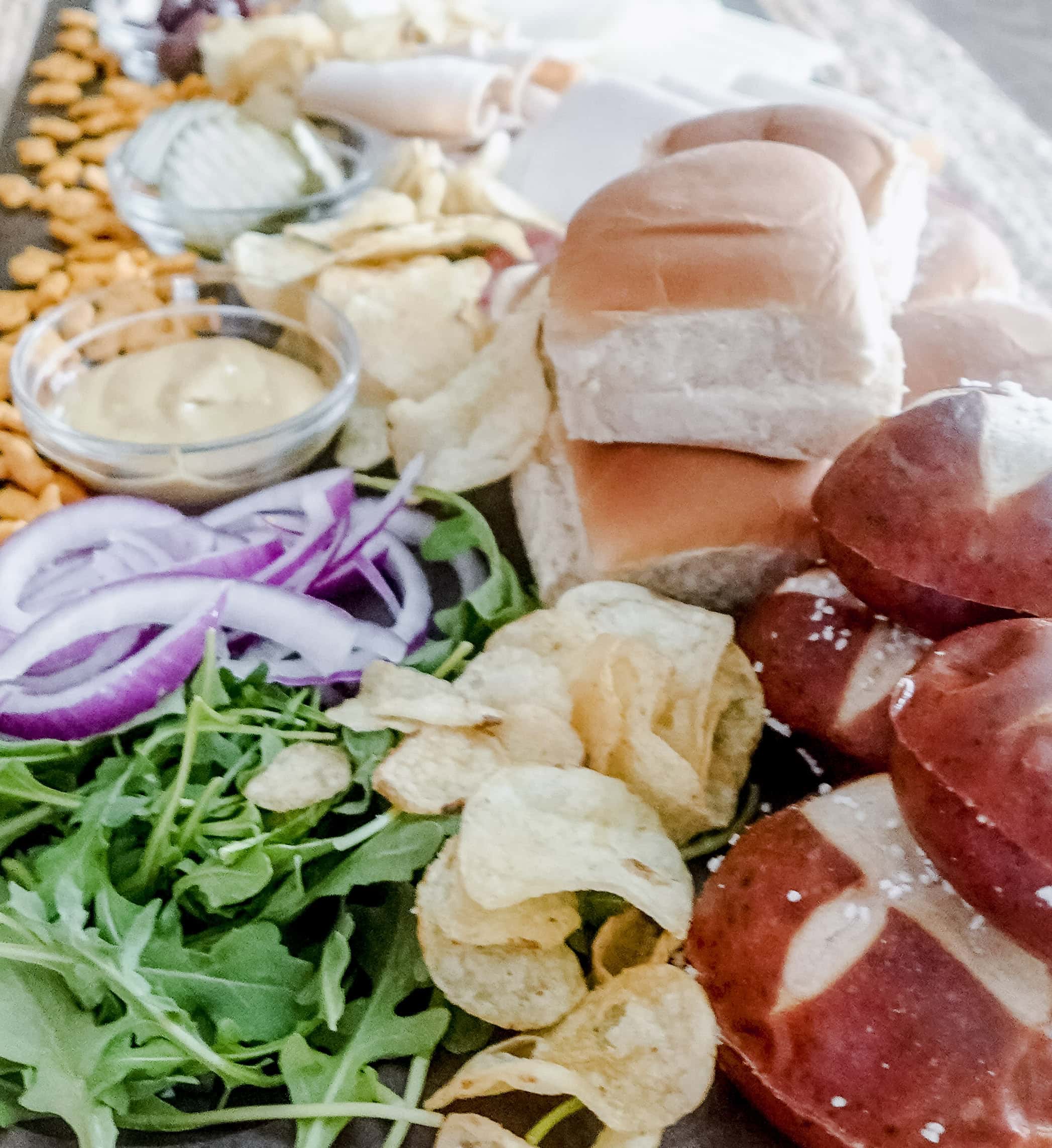 A close-up of a charcuterie board with sandwich rolls, pretzel buns, potato chips, arugula, sliced red onions, mustard, and other assorted snacks.