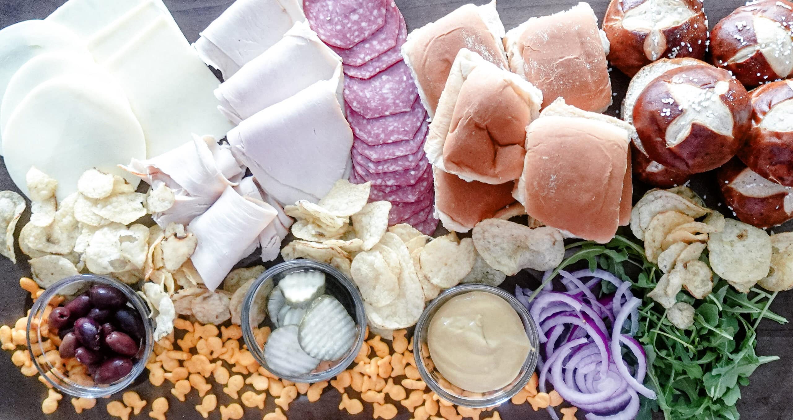 An overhead shot of a sandwich board with sliced cheeses, deli meats, salami, sandwich rolls, pretzel buns, potato chips, goldfish crackers, pickles, olives, mustard, sliced red onions, and fresh arugula on a wooden surface.