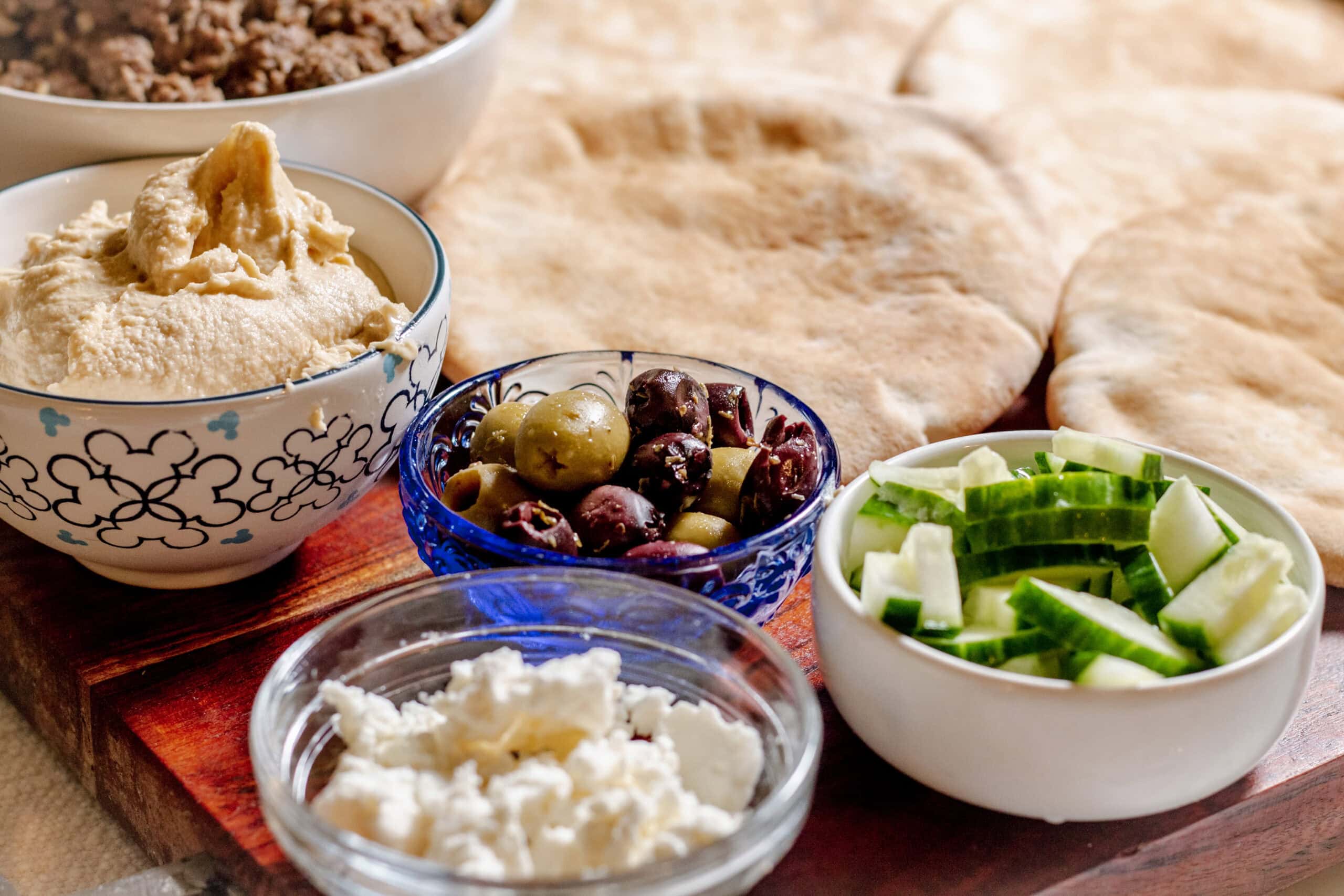A close-up of a Mediterranean spread with bowls of hummus, olives, chopped cucumber, feta cheese, and pita bread on a wooden board.
