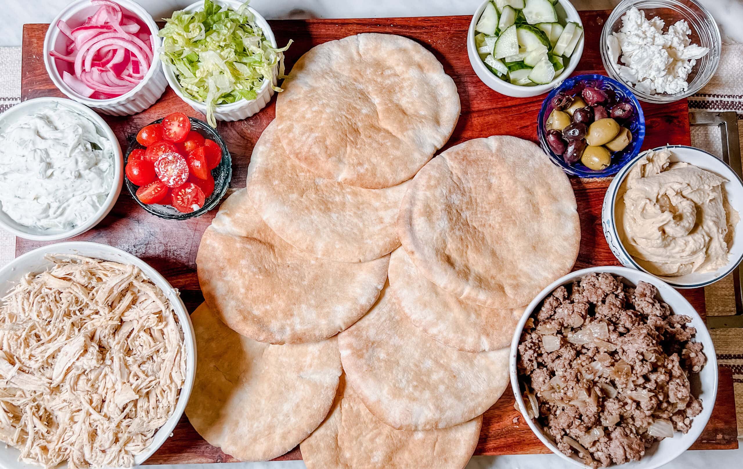 An overhead shot of a wooden board with several pita breads surrounded by bowls of shredded chicken, ground beef, lettuce, pickled onions, cherry tomatoes, cucumber, olives, feta cheese, hummus, and tzatziki sauce.