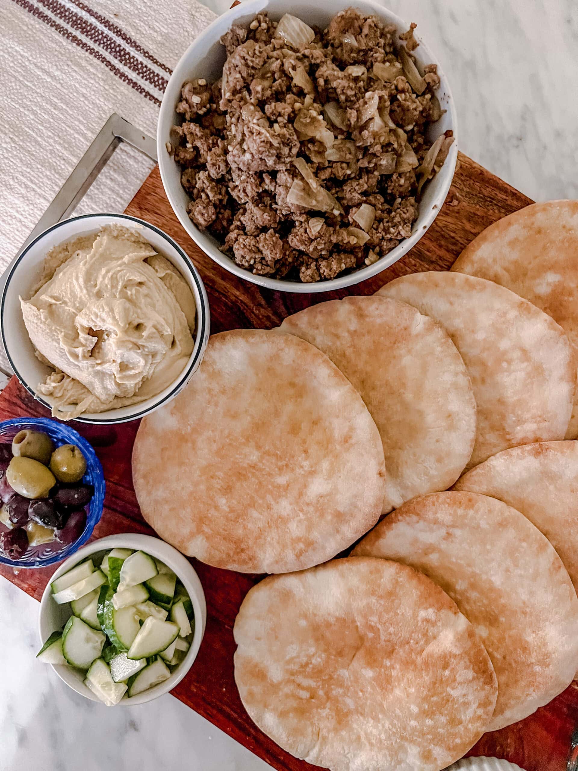 An overhead shot of a wooden board with pita bread, a bowl of cooked ground meat with onions, a bowl of hummus, a small bowl of mixed olives, and a bowl of chopped cucumbers.
