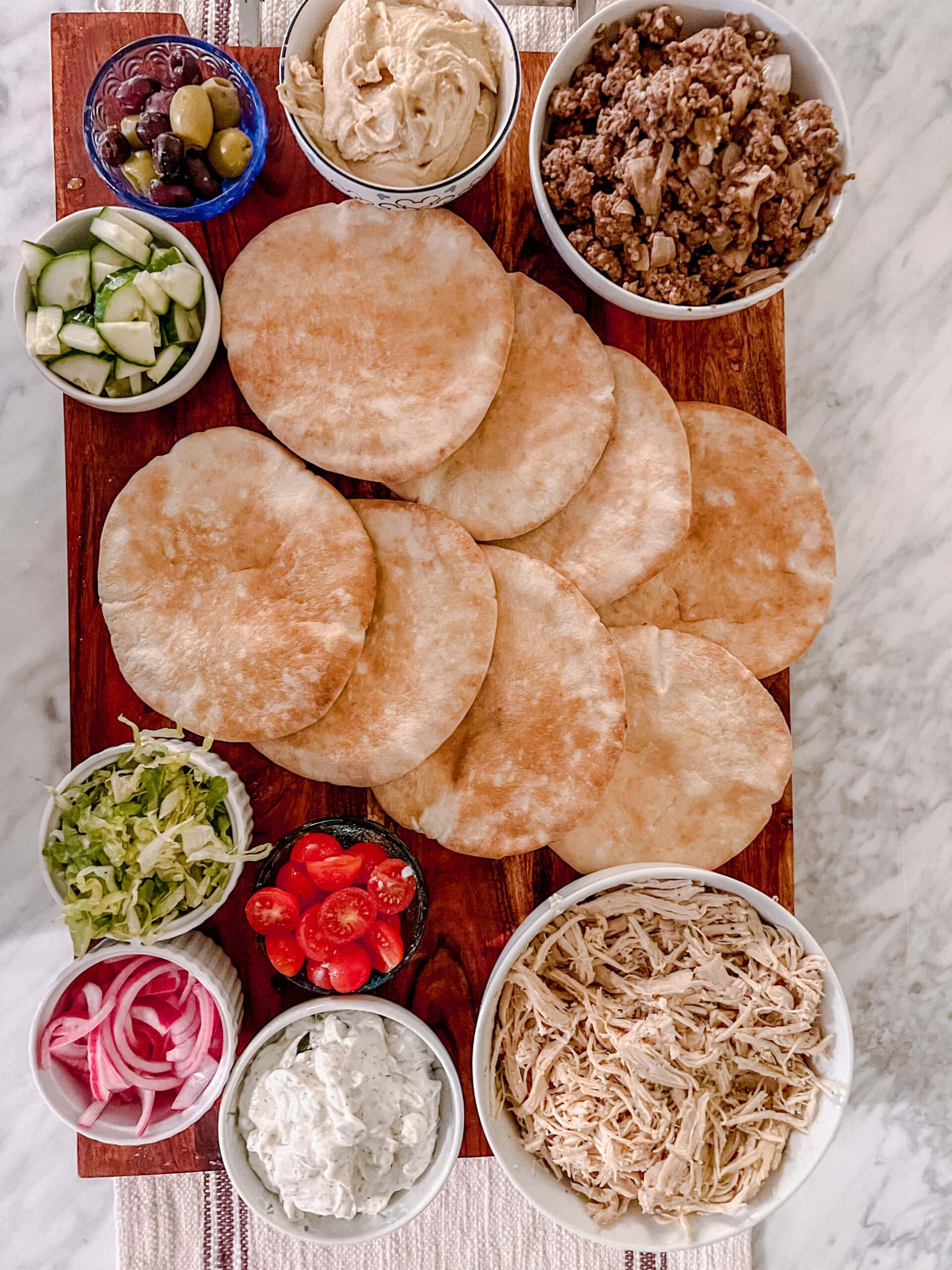An overhead shot of a wooden board with pita bread surrounded by bowls of olives, hummus, cooked ground meat, shredded chicken, lettuce, tomatoes, cucumber, pickled onions, and tzatziki sauce, set on a marble surface.