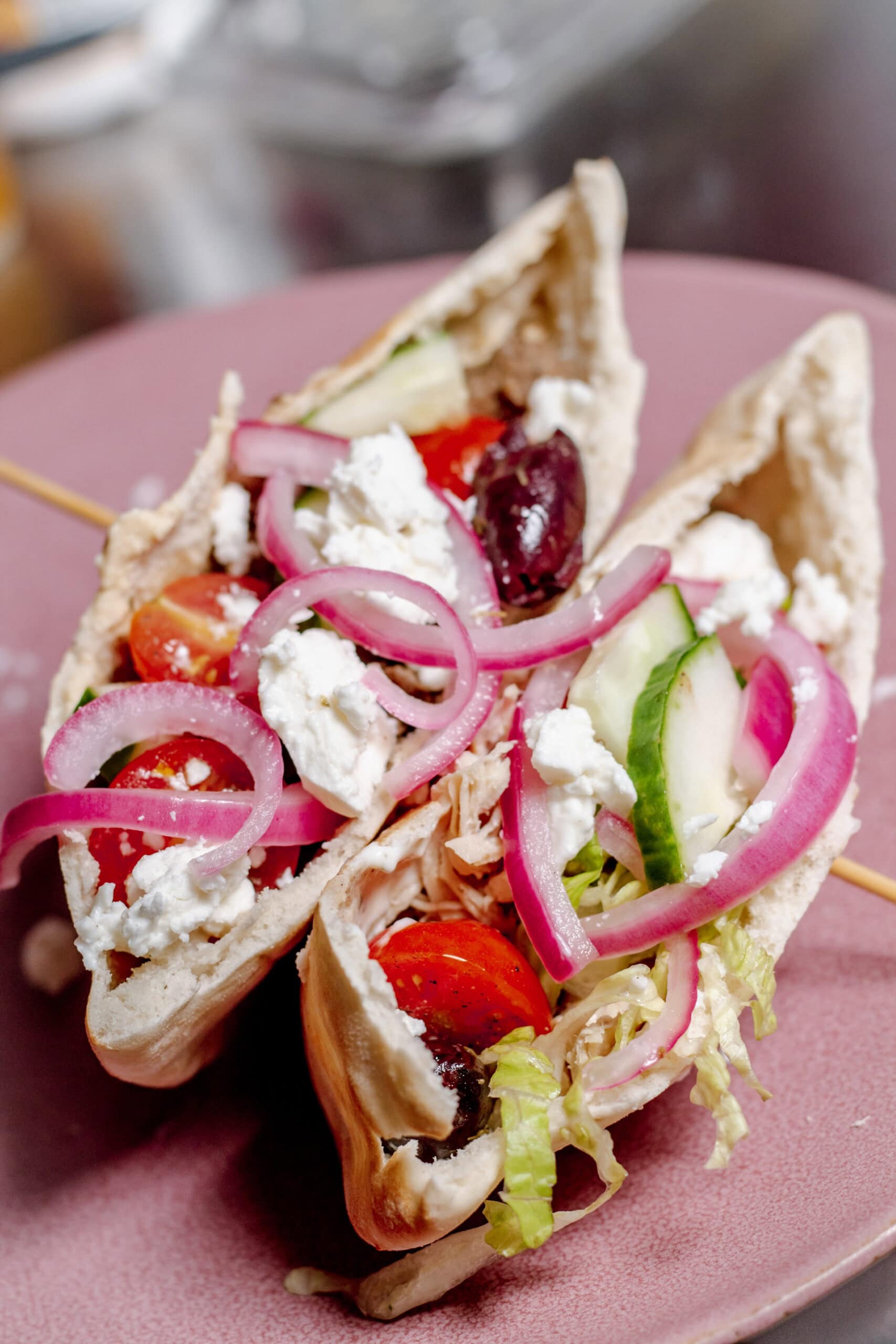 A close-up of two pita pockets filled with shredded lettuce, cherry tomatoes, sliced cucumbers, kalamata olives, pickled red onions, and crumbled feta cheese are served on a pink plate.