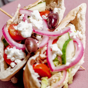 A close-up of two pita pockets filled with lettuce, sliced cucumber, cherry tomatoes, red onion, Kalamata olives, and crumbled feta cheese are served on a pink plate.