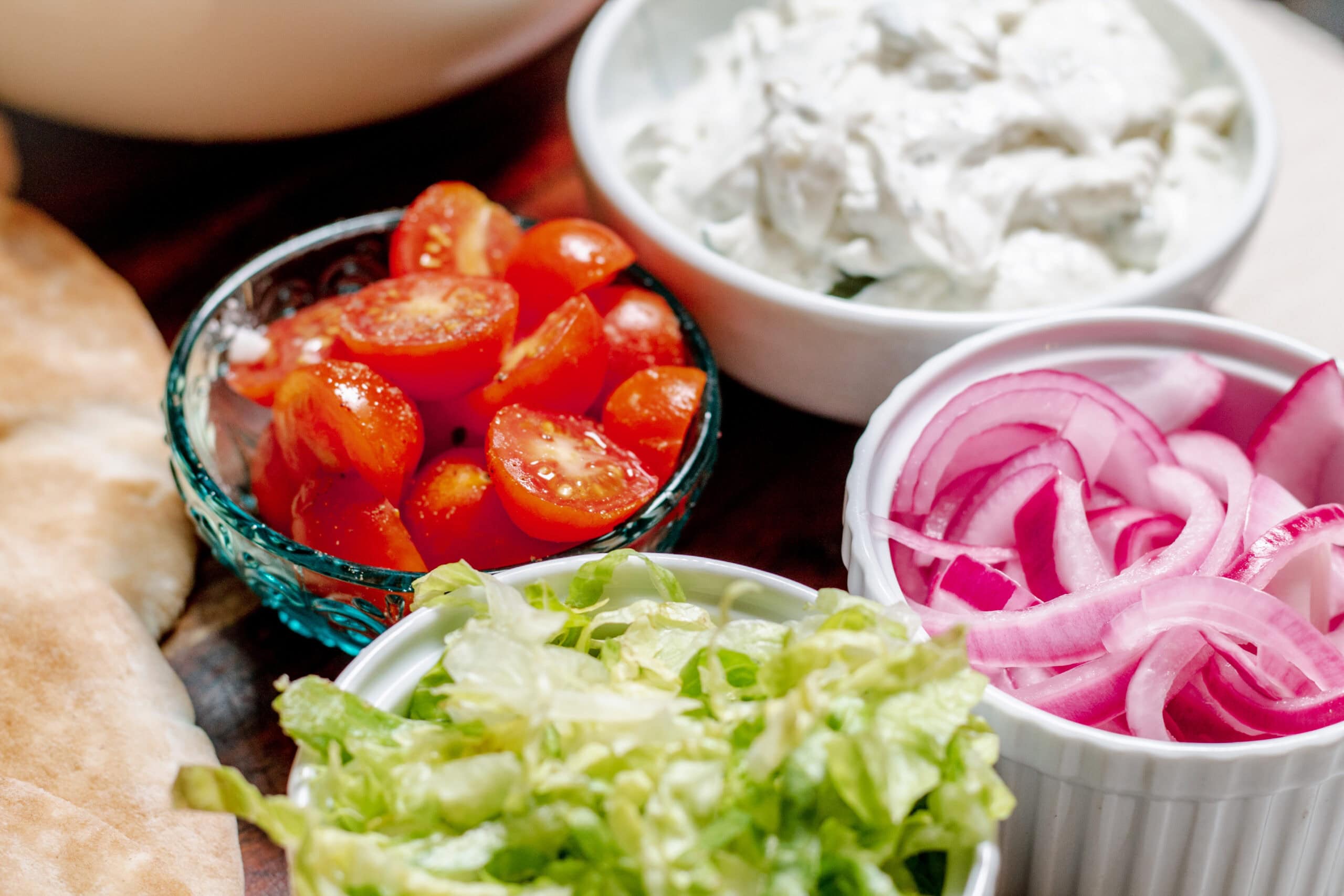 A close-up of assorted fresh ingredients in bowls, including halved cherry tomatoes, sliced red onions, shredded lettuce, and a bowl of creamy dip or spread, all arranged on a wooden surface.
