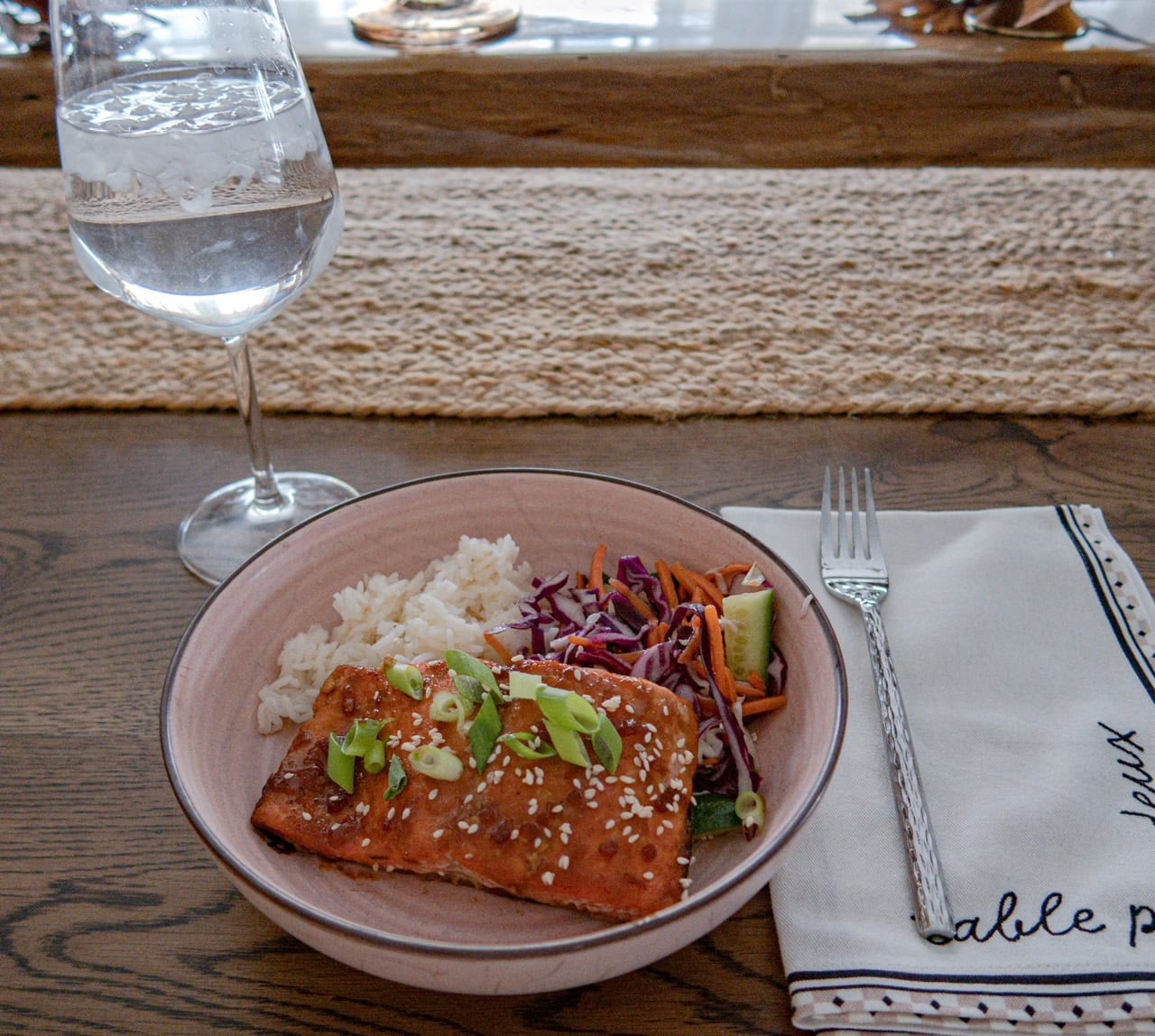 A plate with glazed salmon topped with green onions and sesame seeds, served with white rice and colorful slaw of red cabbage, carrots, and cucumbers, sits next to a fork, napkin, and a stemmed glass of water on a wooden table. There is a woven table runner in the background.