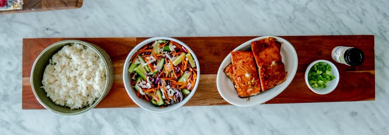 An overhead shot of a wooden board that holds bowls of white rice, colorful mixed salad of cucumbers, carrots and red cabbage, a platter of glazed salmon fillets, a small dish of chopped green onions, and a small bottle of sauce on a white marble surface.
