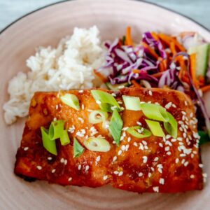 A close-up of a plate with a piece of glazed salmon topped with sliced green onions and sesame seeds, served with white rice and a colorful side of shredded cabbage, carrots, and cucumber salad.