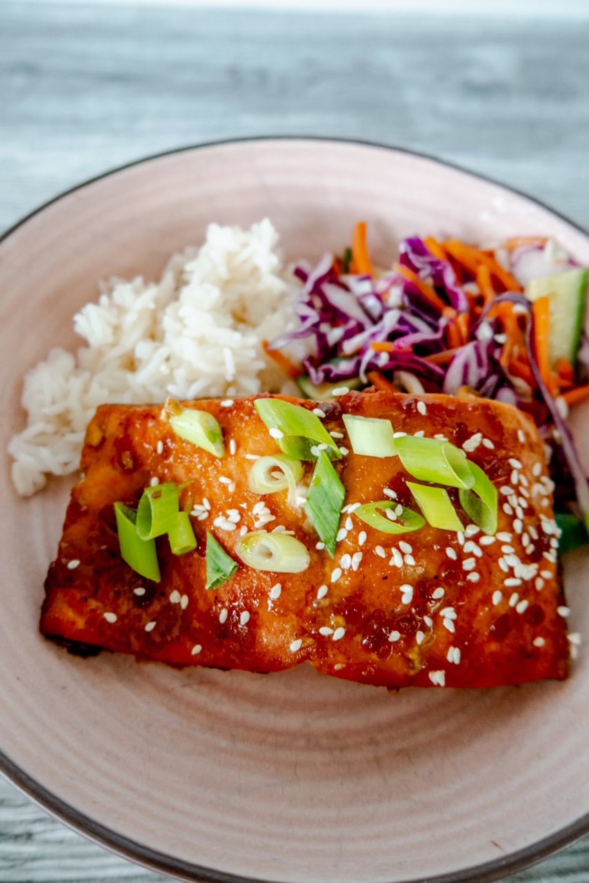 A close-up of a plate with a piece of glazed salmon topped with sliced green onions and sesame seeds, served with white rice and a colorful side of shredded cabbage, carrots, and cucumber salad.