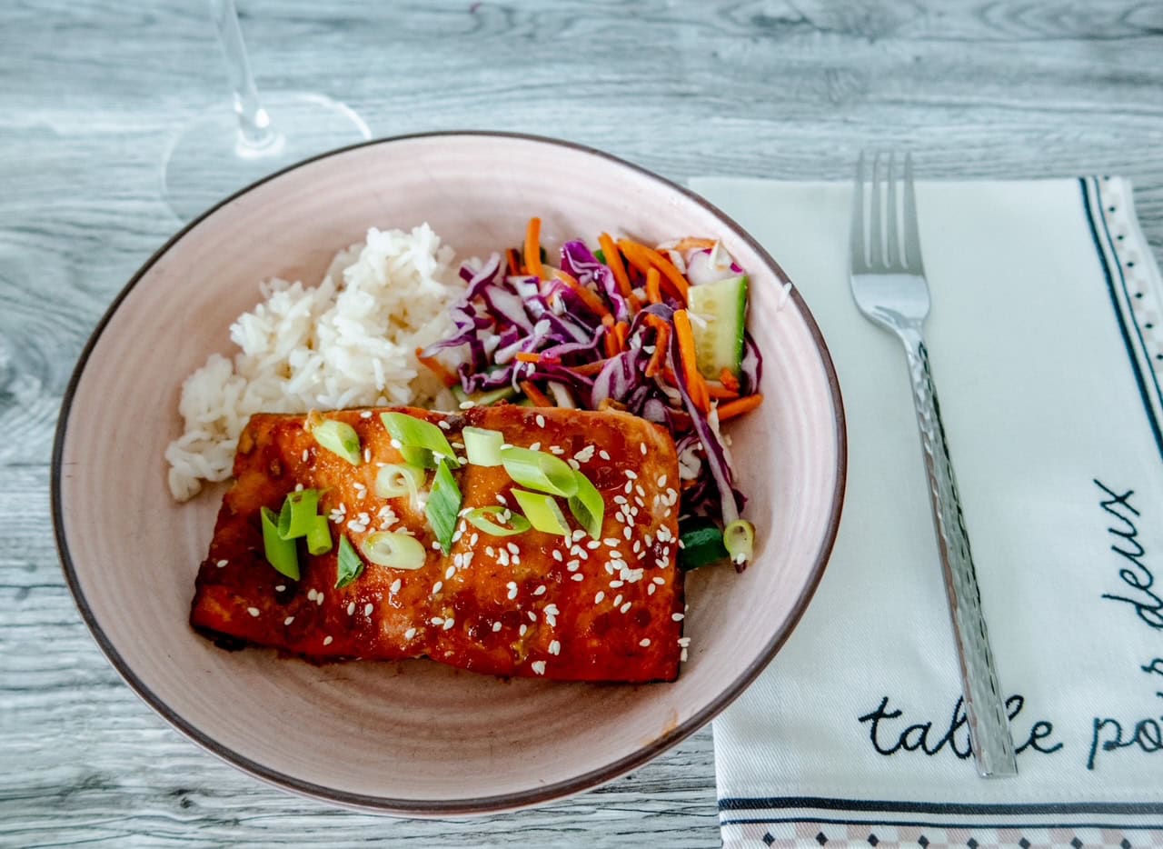 A plate with glazed salmon topped with green onions and sesame seeds, served with white rice and a colorful slaw of red cabbage, carrots, and cucumber. A fork rests on a napkin beside the plate and a wine glass can be seen sitting on the gray wooden table above the plate.