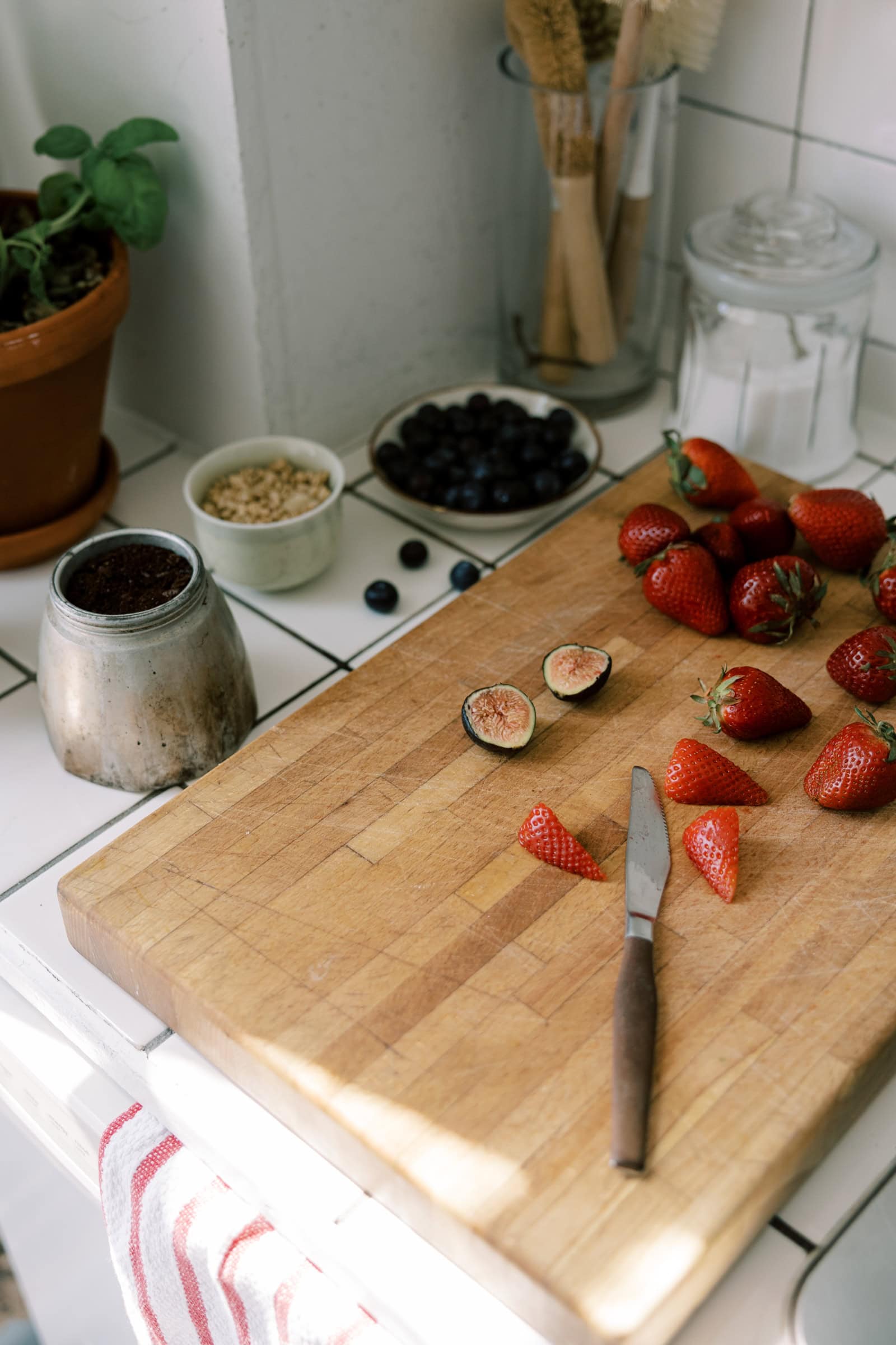 A wooden cutting board on a kitchen counter with sliced strawberries, a fig, and a knife. Nearby are a potted plant, bowl of blueberries, jar of sugar, the bottom of a moka pot, and a container with kitchen utensils.