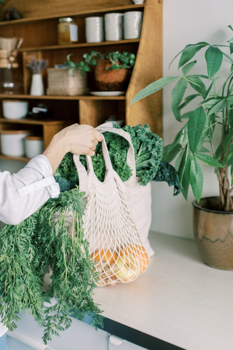 A person holds reusable bags filled with fresh leafy greens, carrots, and oranges in a bright kitchen with a potted plant and wooden shelves in the background.