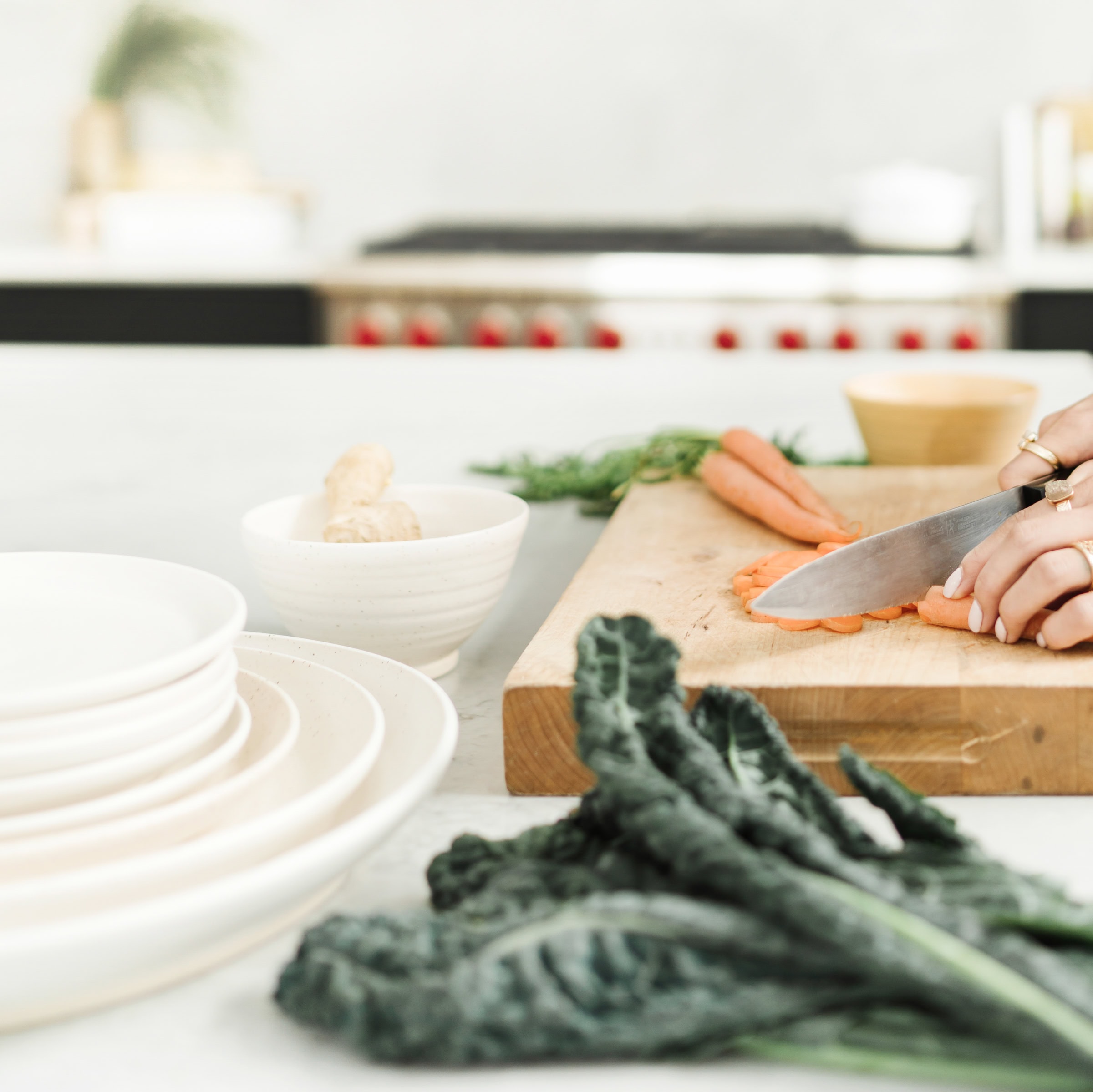 A person slices carrots on a wooden cutting board in a bright kitchen. Plates, ginger, and leafy greens are visible on the counter nearby.