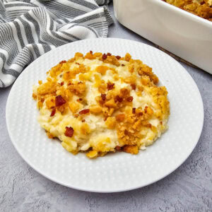 A serving of creamy casserole topped with a golden, crunchy crumb layer sits on a white plate, next to a striped kitchen towel and a baking dish on a light gray surface.