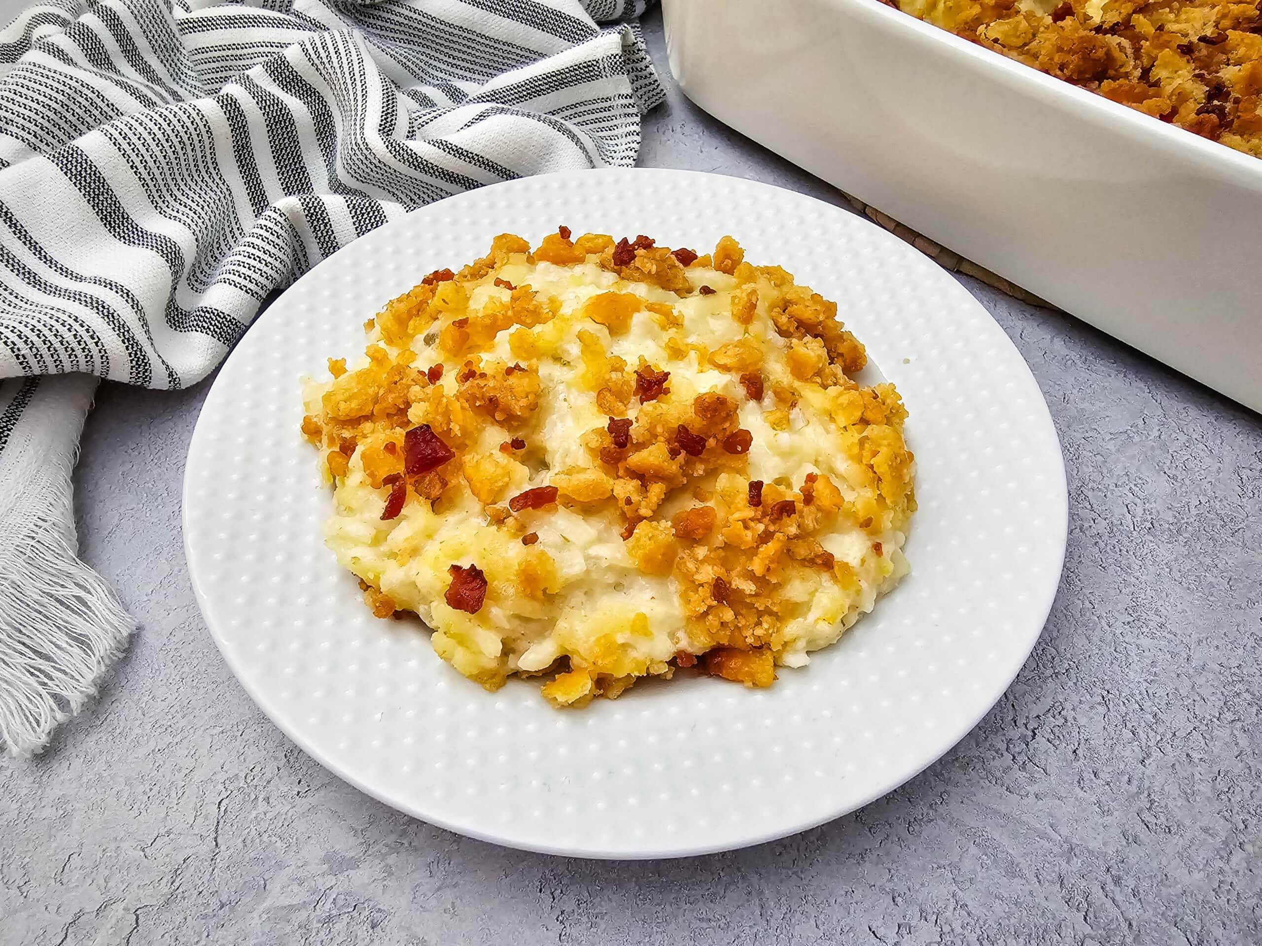 A serving of creamy casserole topped with a golden, crunchy crumb layer sits on a white plate, next to a striped kitchen towel and a baking dish on a light gray surface.