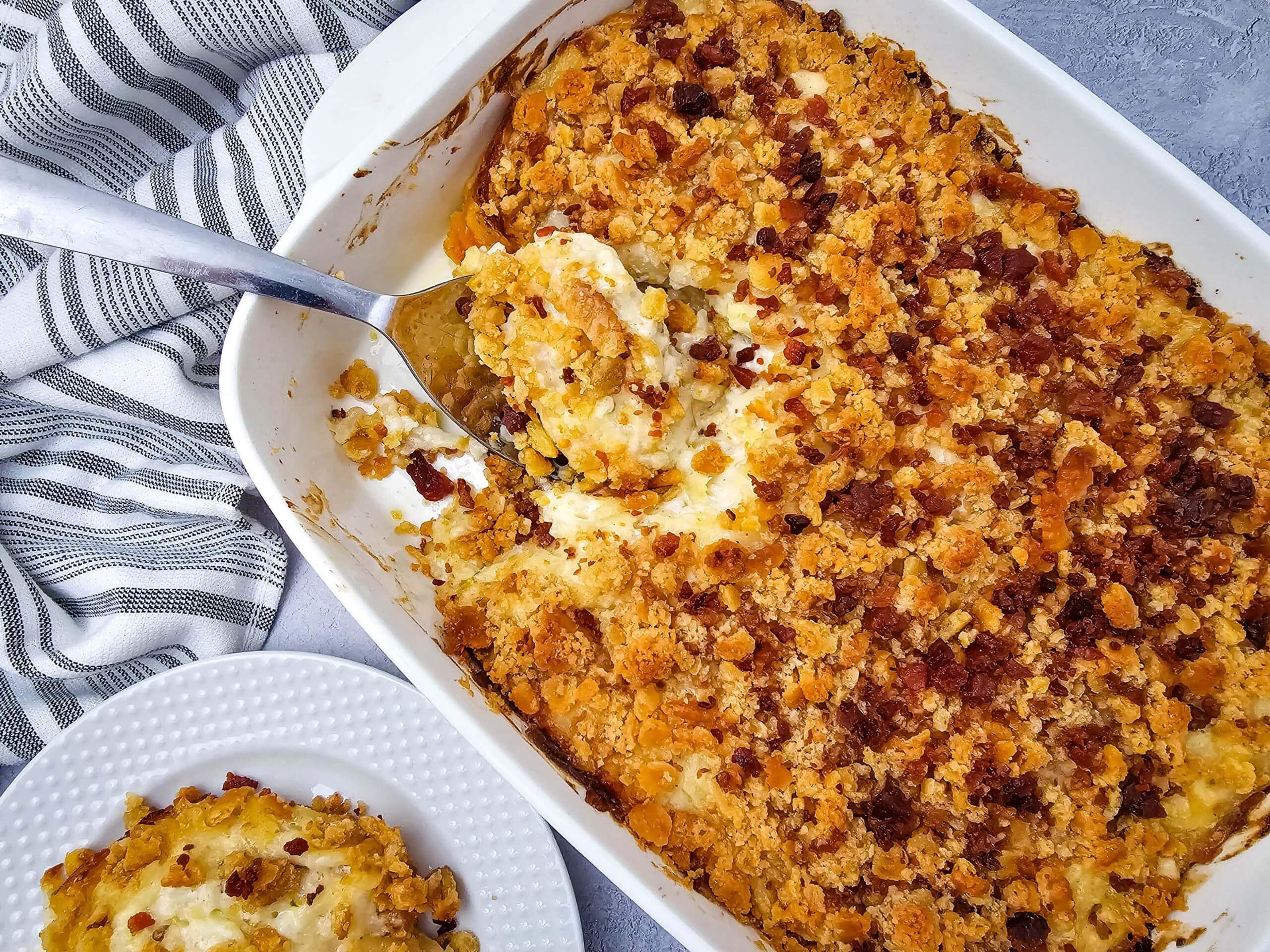 A casserole dish filled with a golden, cracker-topped baked potato casserole, with a serving spoon lifting out a portion. A plate with a serving of the casserole sits nearby on a gray surface next to a striped cloth.