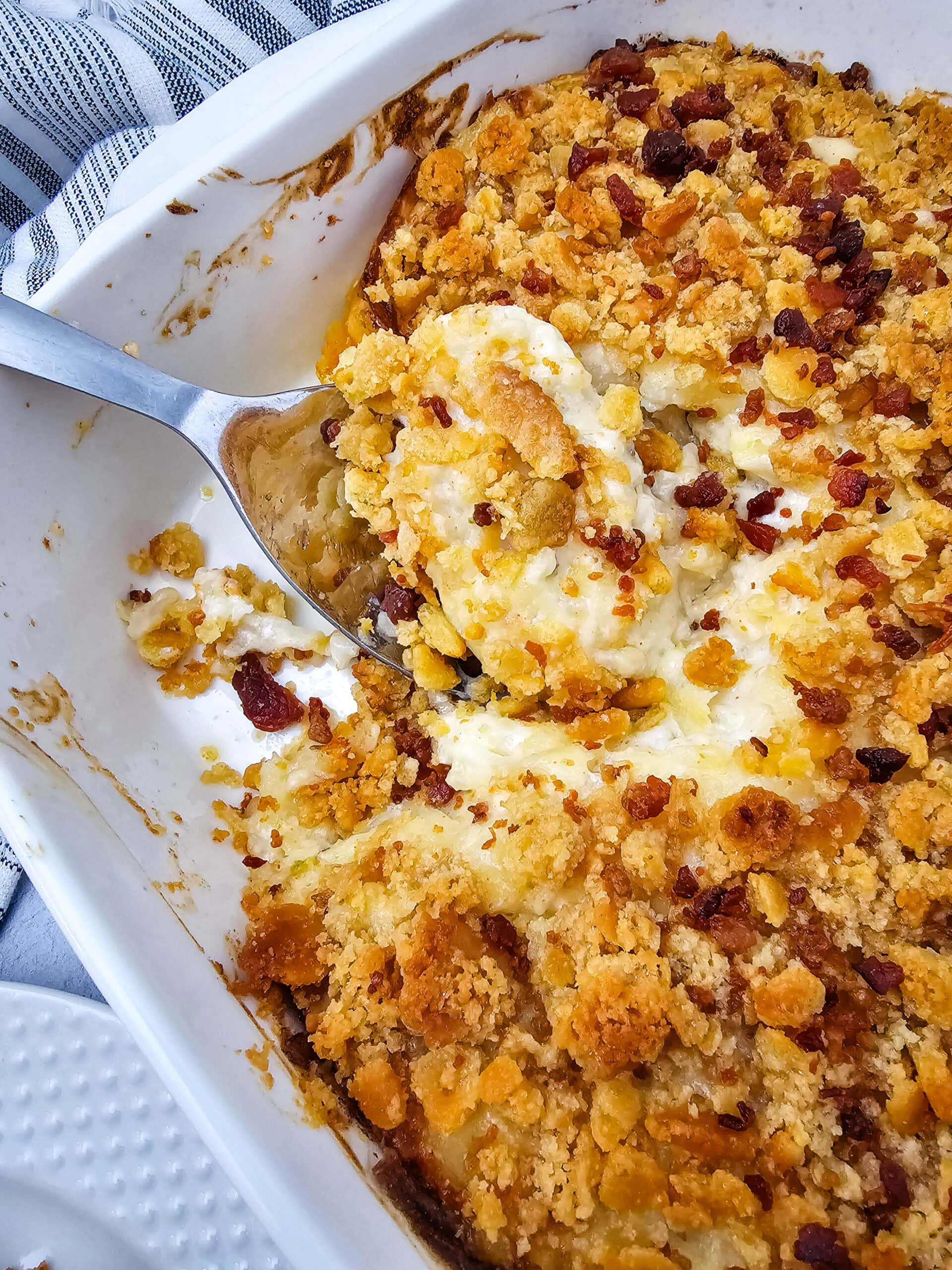 A close-up of a baked potato casserole in a white dish, topped with a golden, crispy cracker and bacon topping. A metal serving spoon is lifting out a portion, revealing a creamy filling underneath.
