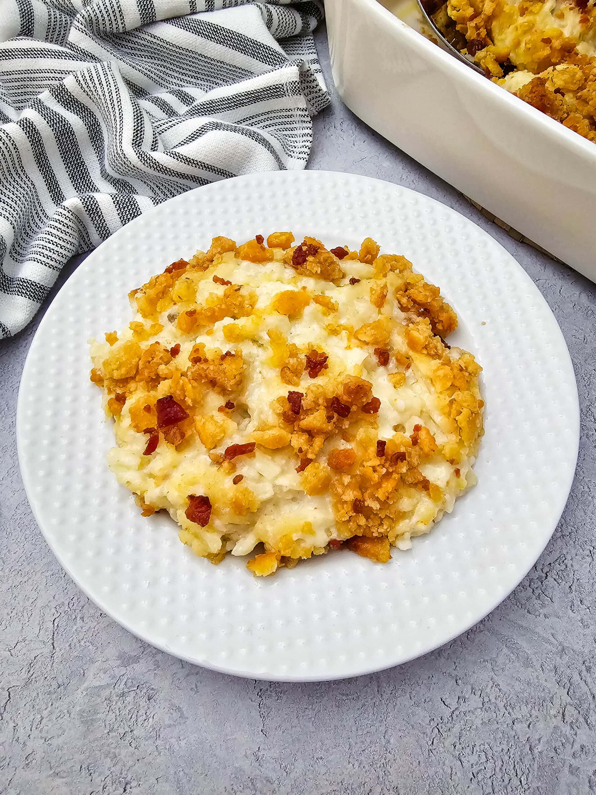 A serving of cheesy potato casserole topped with a golden, crunchy breadcrumb crust and bacon is plated on a white dish, with a striped kitchen towel and casserole dish visible in the background.