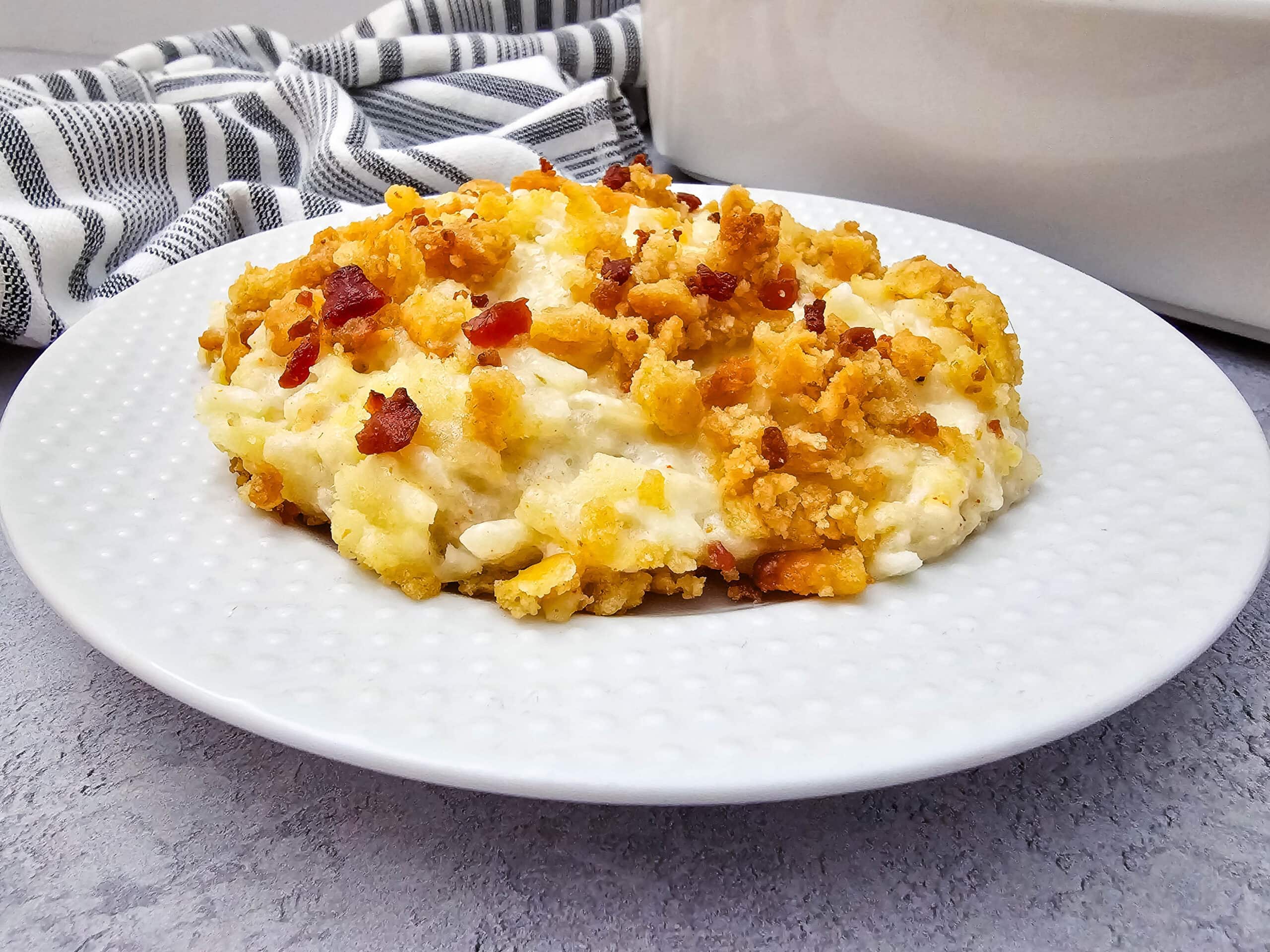 A serving of creamy potato casserole topped with golden, crumbly breadcrumbs and bits of bacon is on a white plate, with a striped kitchen towel and a white dish in the background.
