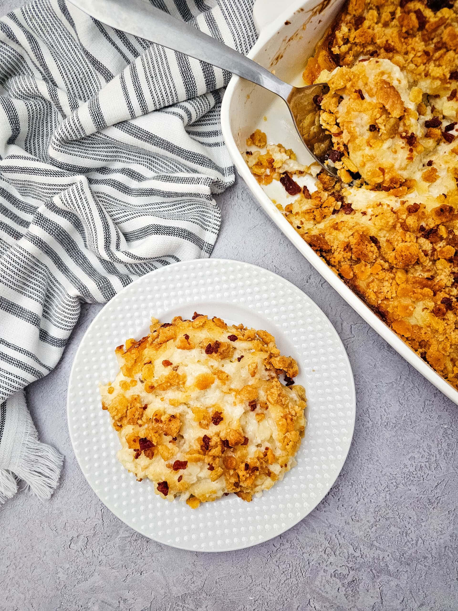 A round serving of baked potato casserole with a golden cracker topping sits on a white plate beside a striped kitchen towel, with the rest of the casserole in a white baking dish and a serving spoon nearby.
