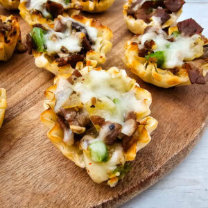 A close-up of mini phyllo pastry cups filled with diced steak, green bell peppers, mushrooms, and melted cheese, arranged on a wooden serving board.