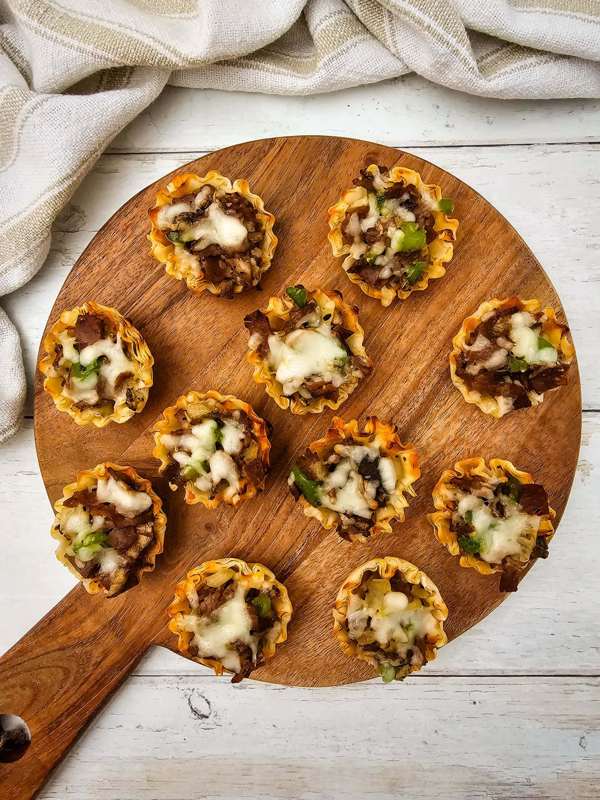 An overhead shot of mini phyllo pastry cups filled with a mixture of beef, vegetables, and melted cheese are arranged on a round wooden board atop a white surface, with a beige cloth in the background.