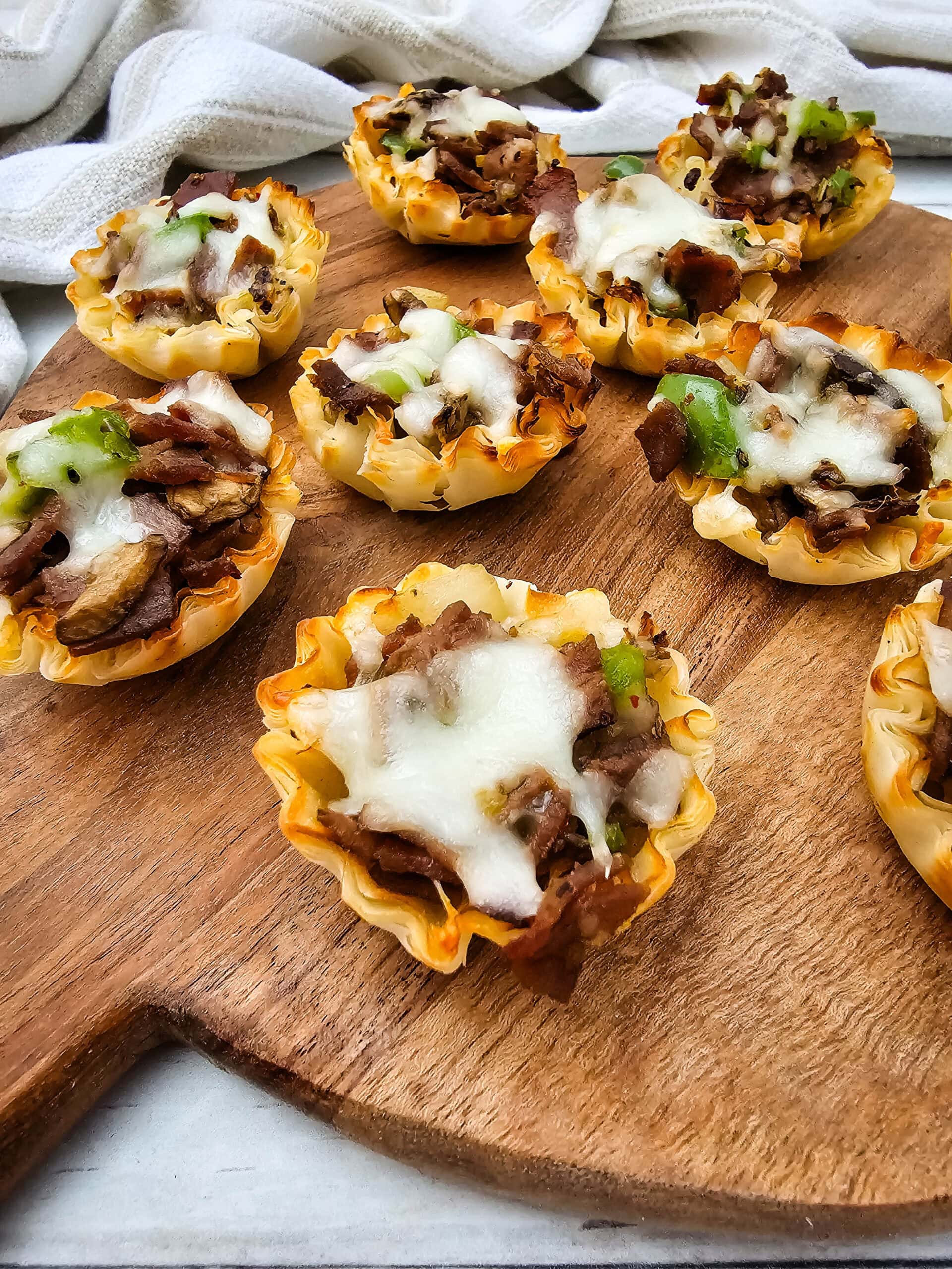 Mini phyllo pastry cups filled with beef, green bell peppers, and melted cheese, arranged on a wooden serving board. A light-colored cloth is in the background.