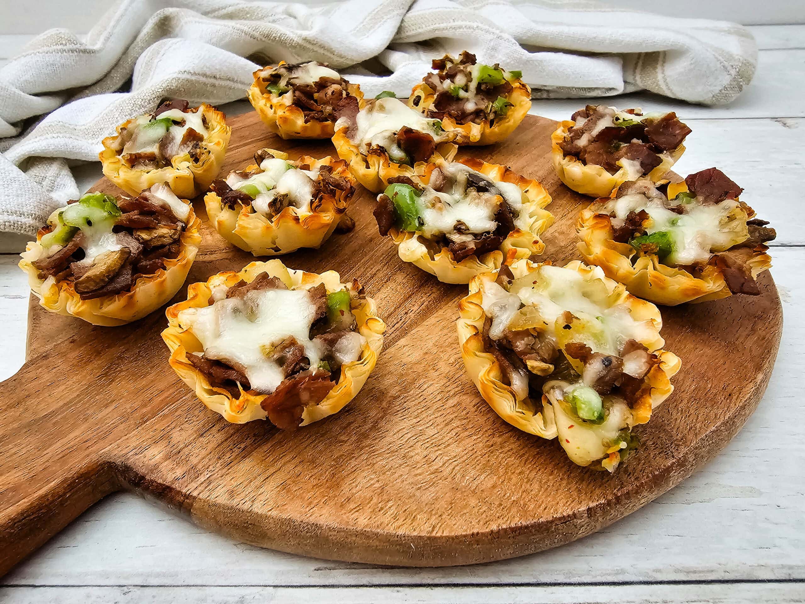Mini phyllo pastry cups filled with diced steak, green peppers, and melted cheese are arranged on a round wooden serving board, with a white cloth in the background.