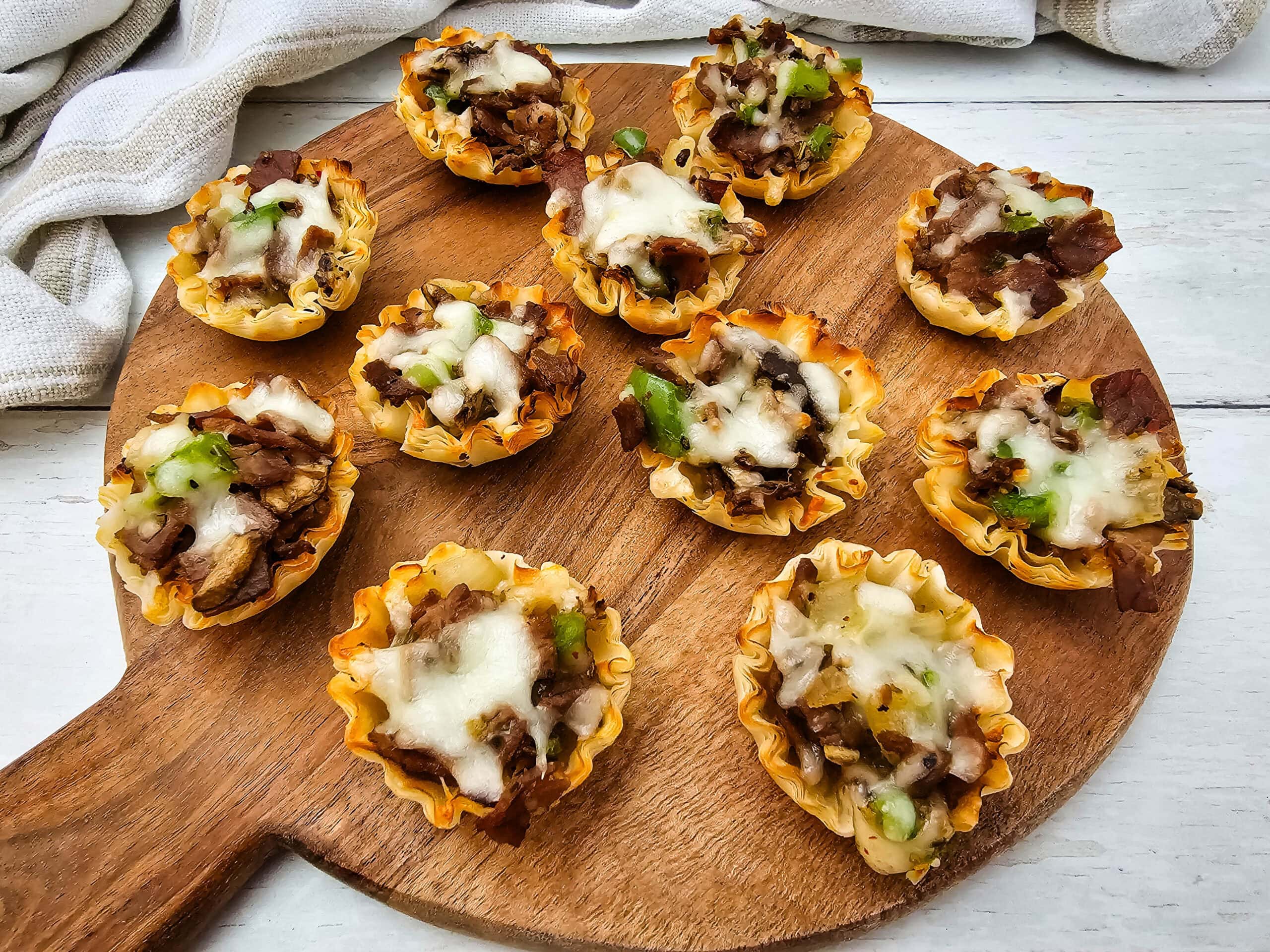 Mini phyllo pastry cups filled with seasoned beef, vegetables, and melted cheese are arranged on a round wooden serving board, with a white cloth in the background.