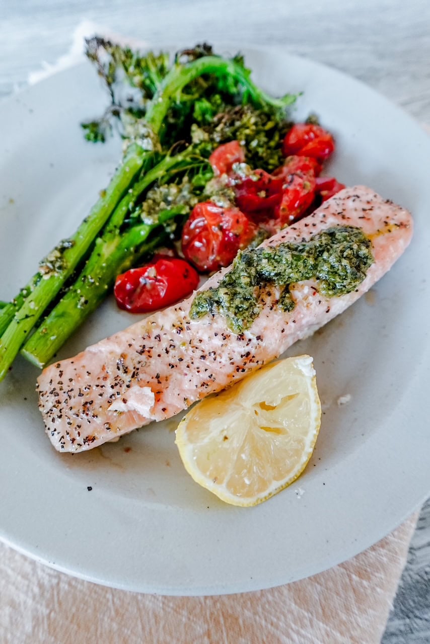 A plate with a piece of baked salmon topped with green herb sauce, served with broccolini, roasted cherry tomatoes, and a lemon wedge.